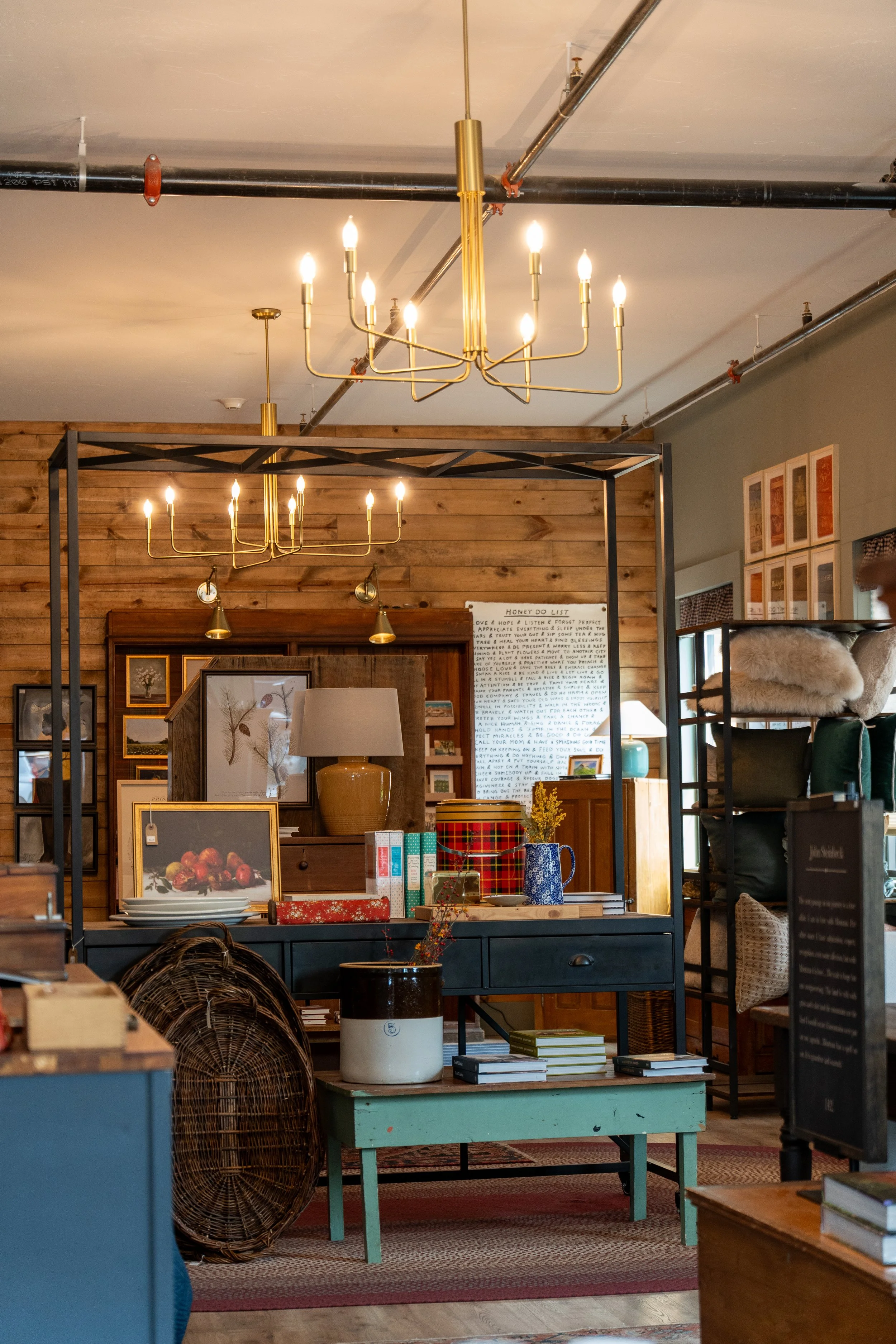 Interior view of a home decor or antique store with warm lighting. Two golden chandeliers hang from the ceiling, illuminating shelves filled with various decorative items, framed art, and plants. The space features wooden accents, posters on the wall, and a variety of furniture and decor items on display.