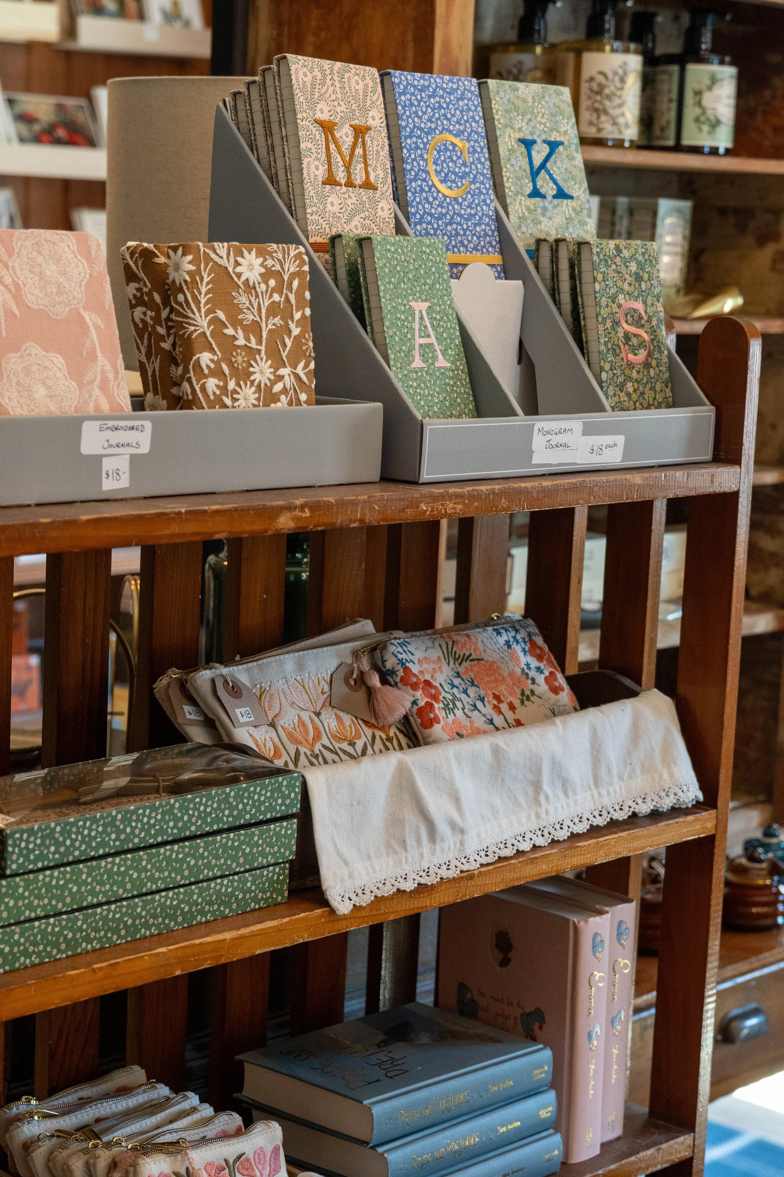 Display shelf with decorative journals and notebooks, some with floral and botanical cover designs, including alphabet letters and patterns, in a store setting.