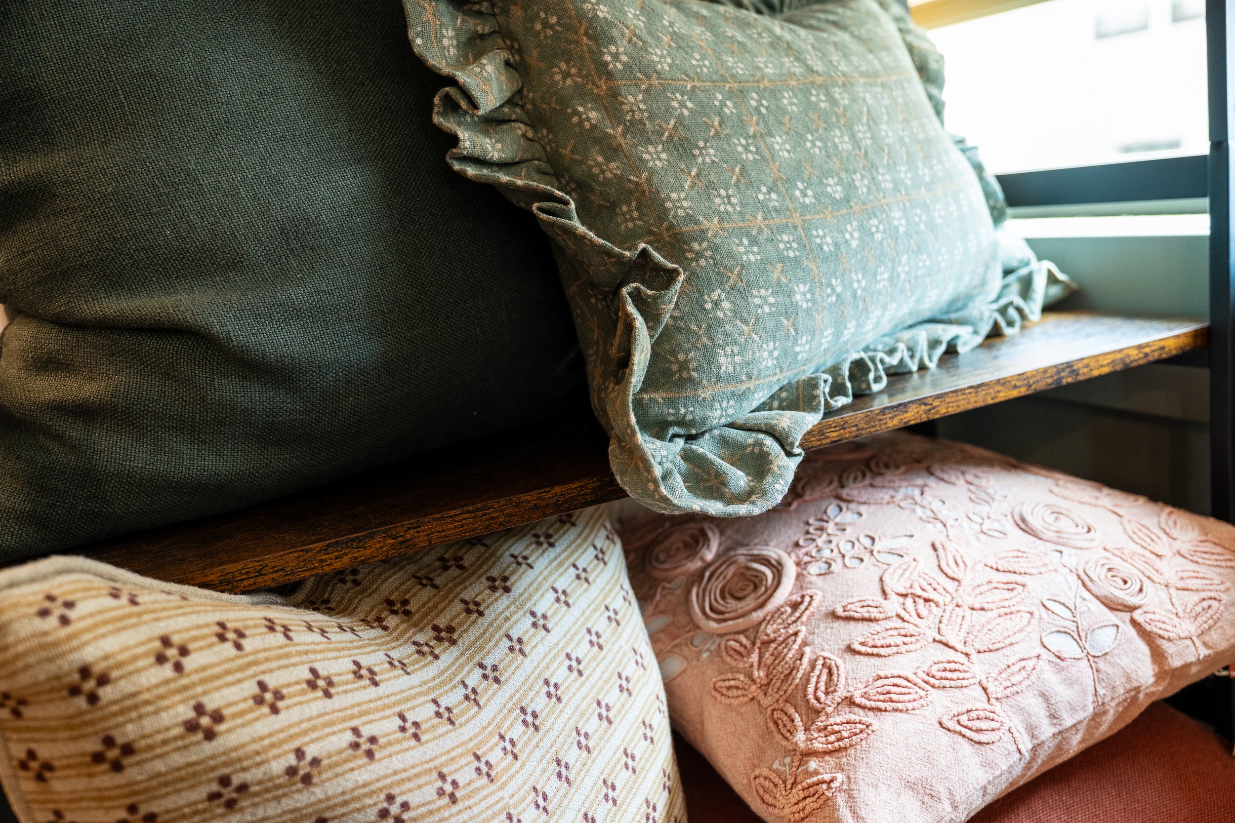 Assorted decorative pillows on a wooden shelf near a window.