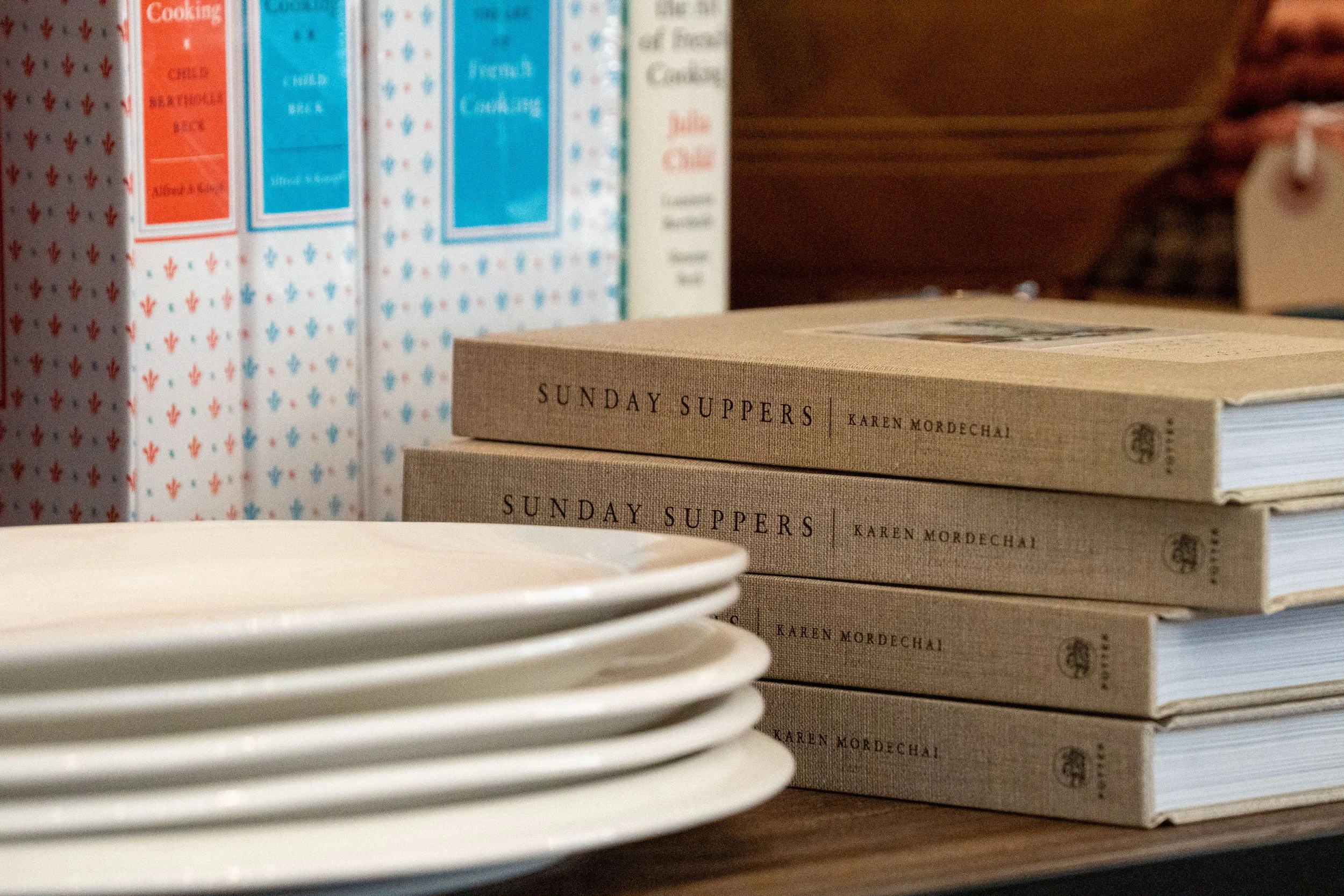 Stack of beige books titled "Sunday Suppers" by Karen Mordechai, lined up on a shelf next to a row of white plates, with patterned boxes or binders in the background.