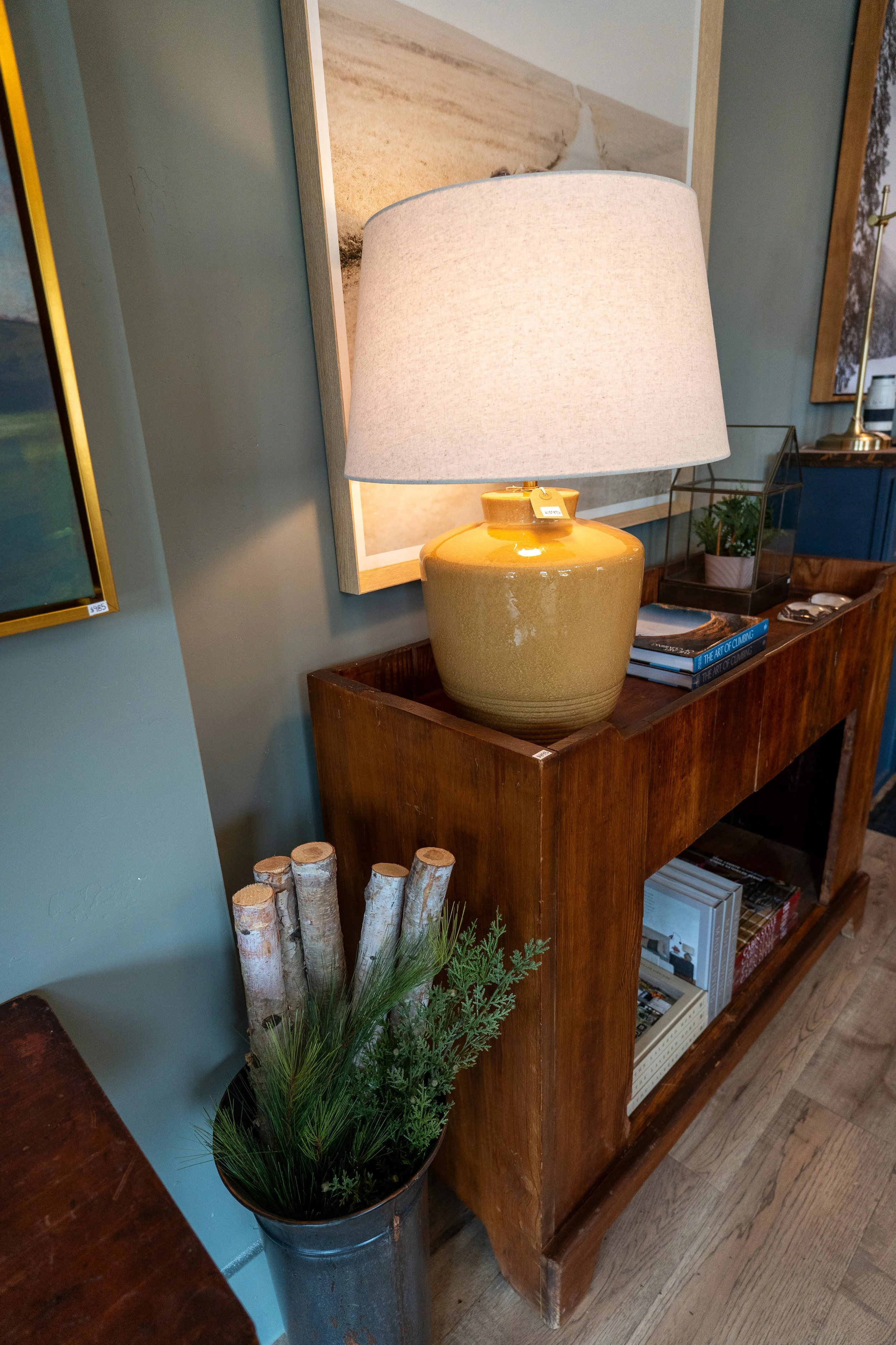 A wooden bookshelf with decorative items and books, a table lamp with a beige shade and yellow ceramic base, a small plant in a glass terrarium, and a black and white book titled 'The Art of Climbing'. There is also a basket of logs and a plant in a 