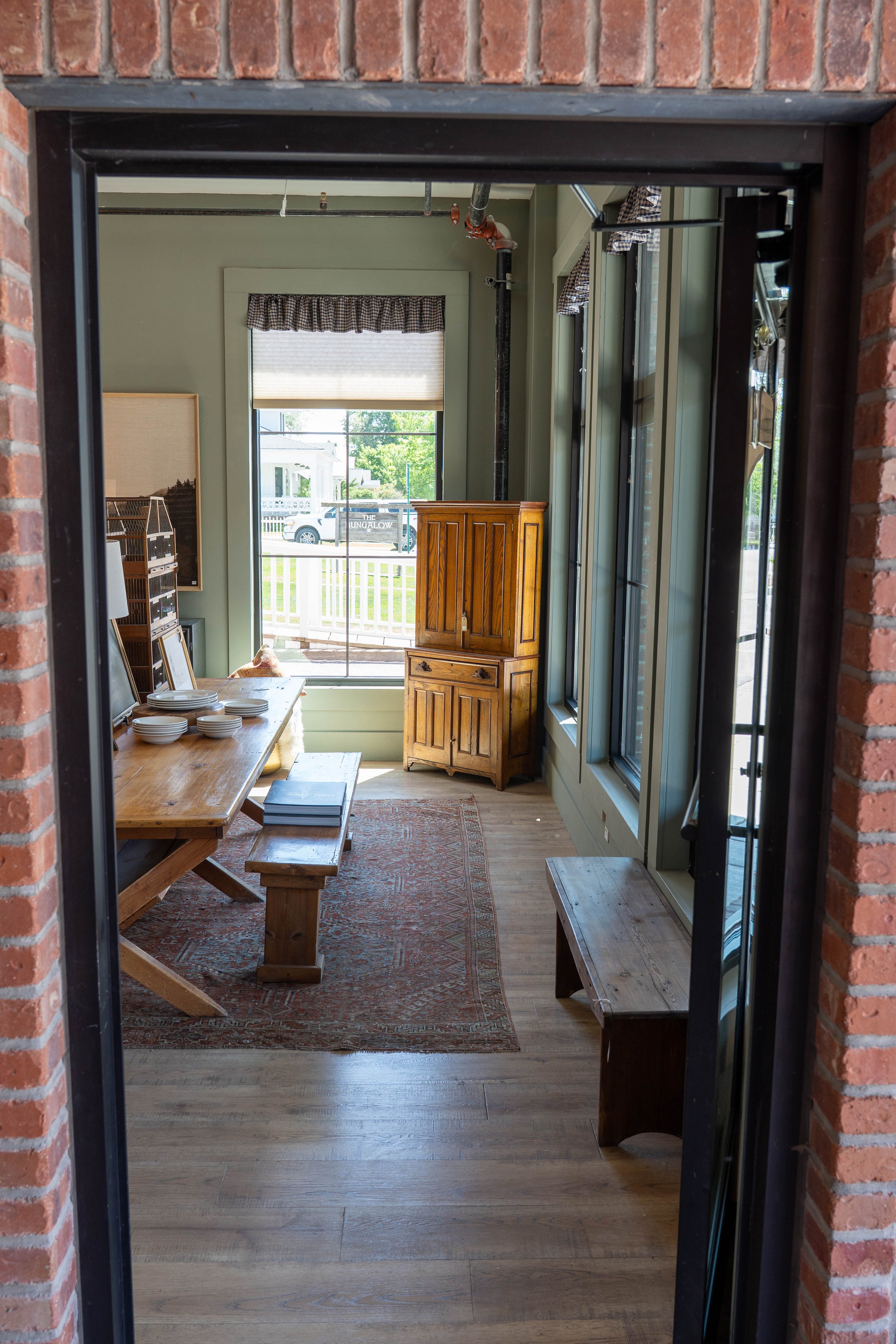 View of an interior room with a wooden dining table, chairs, a wooden cabinet, a sliding glass door leading outside, and large windows with checkered curtains.