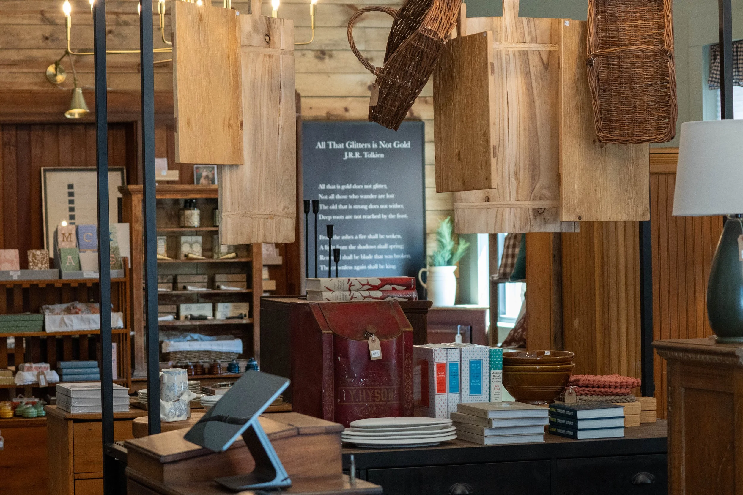 Interior view of a rustic shop display with wooden shelves and wicker baskets, items including books, ceramics, and decorative objects, and a black sign with white text in the background.