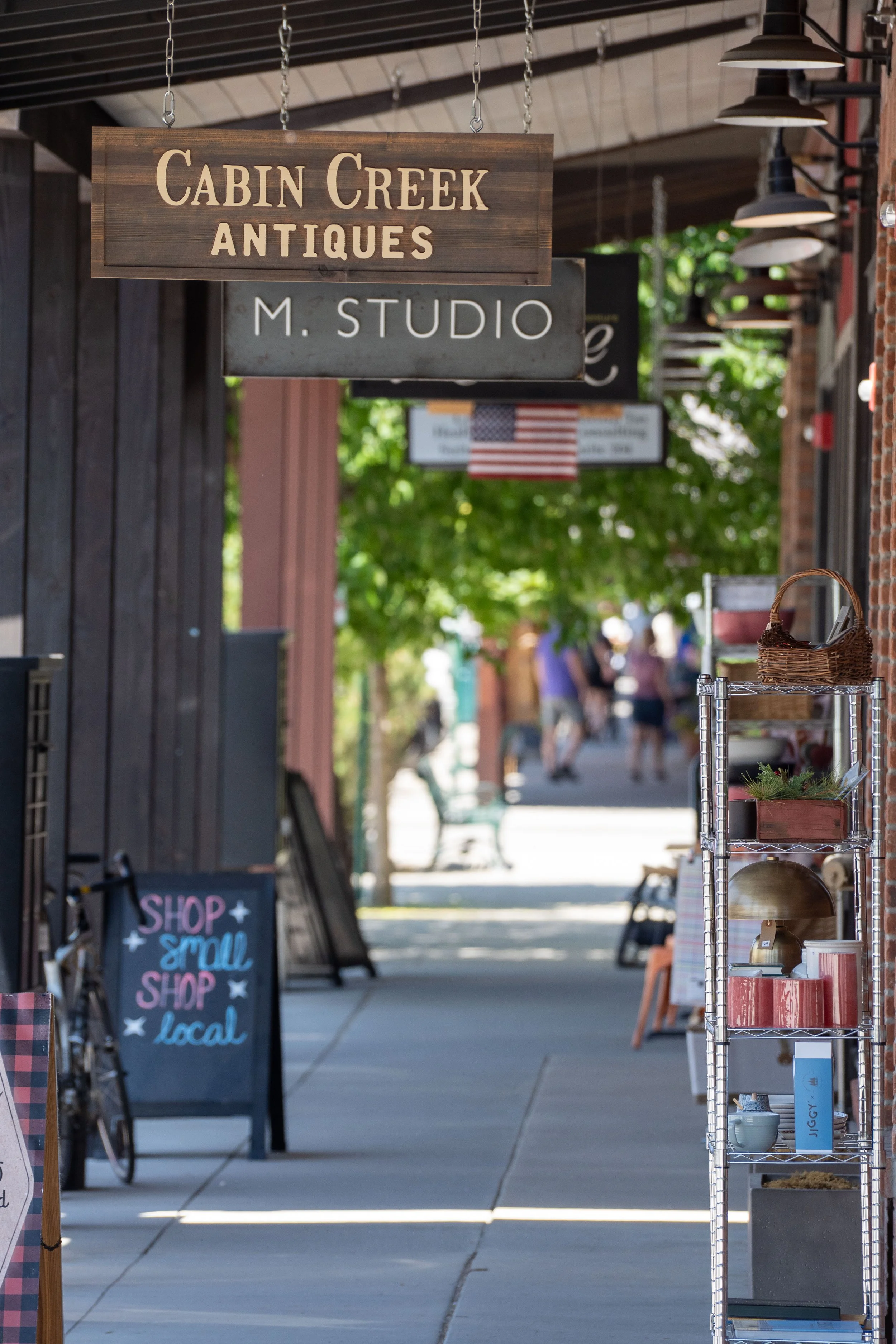A sidewalk storefront with signs for Cabin Creek Antiques and M. Studio, outdoor shelving with pottery and decorative items, and a chalkboard sign that says "Shop small shop local." There are blurry pedestrians and green trees in the background.