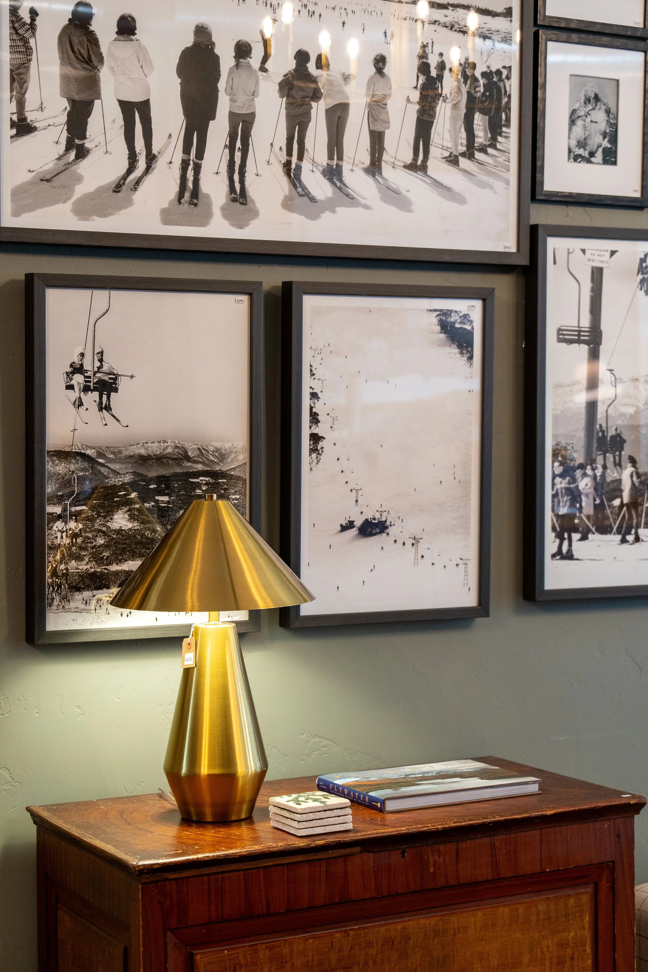 A wooden table with a large, gold-colored, cone-shaped lamp, a book titled 'Fly Water,' and a small stack of coasters. Behind the table is a wall displaying framed black-and-white photographs of skiing, snowboarding, and outdoor winter scenes. The ph