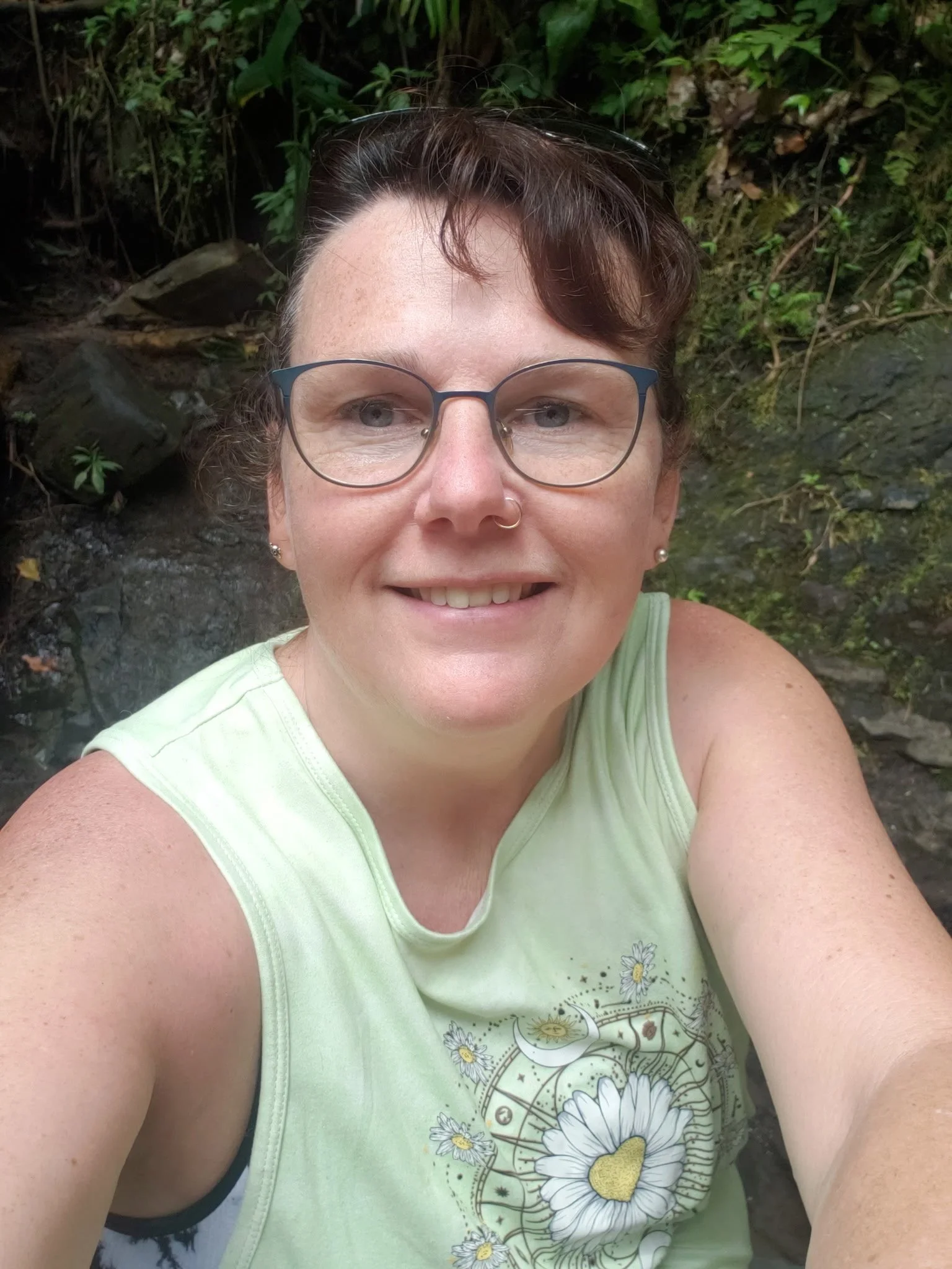 A person with short dark brown hair, glasses, and a nose ring, smiling outdoors amidst greenery and rocks.