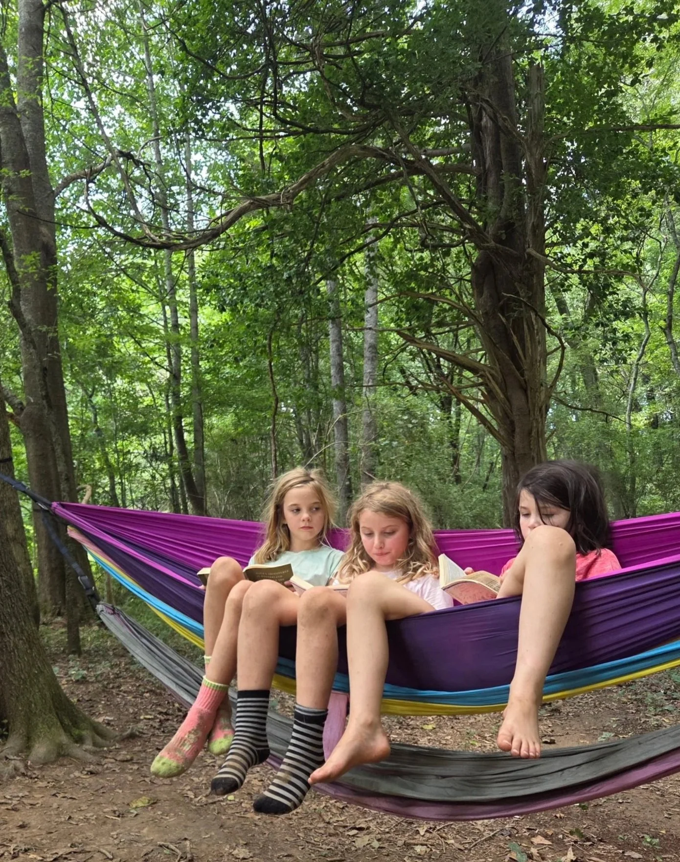 Three girls sitting together in a rainbow-colored hammock in a wooded area, reading books.