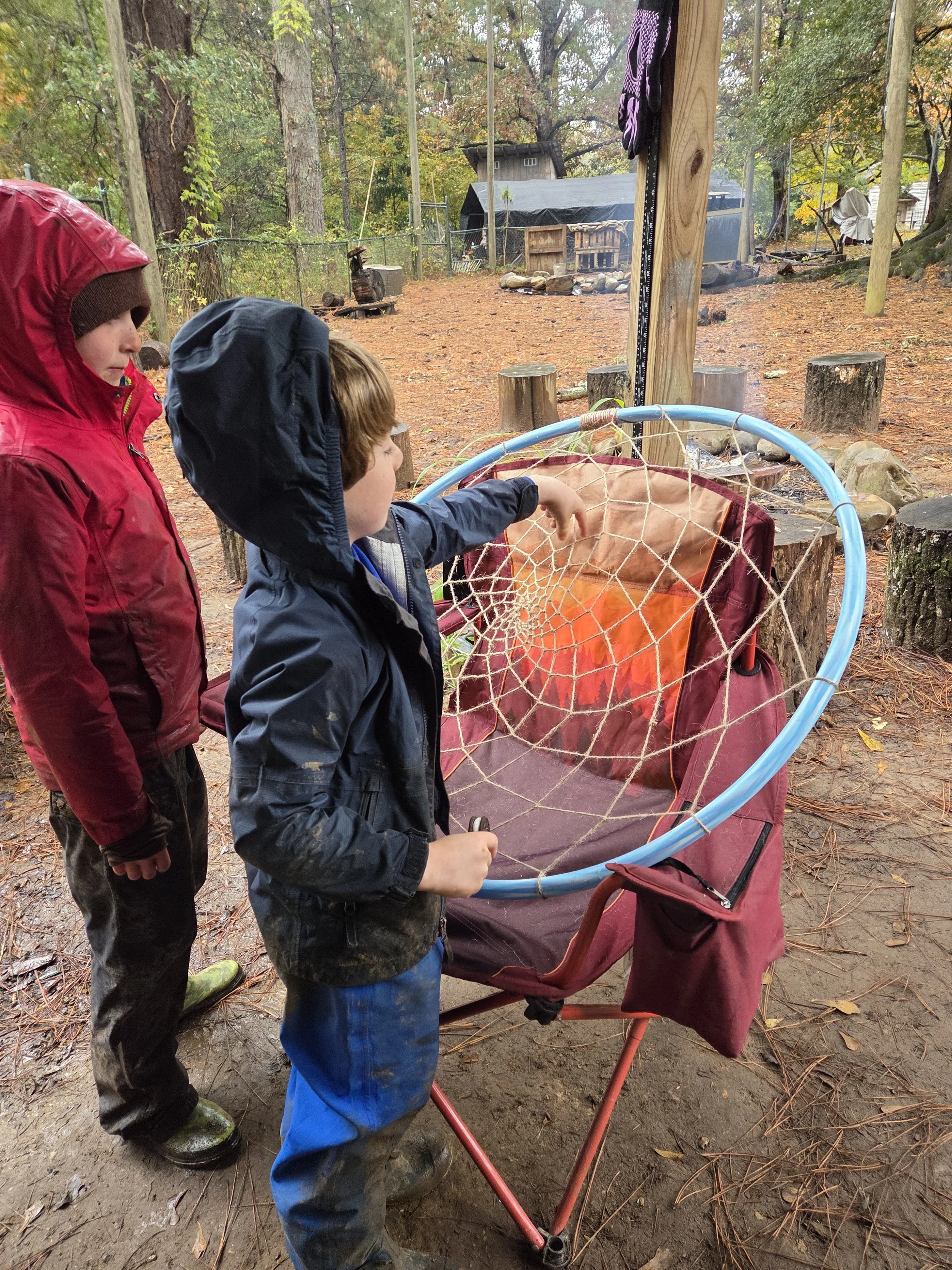 Two children outdoors in a wooded area, one wearing a red jacket with a hood and the other a blue jacket, standing near a red stroller with a net-like cover, and a background of trees, logs, and a shelter.