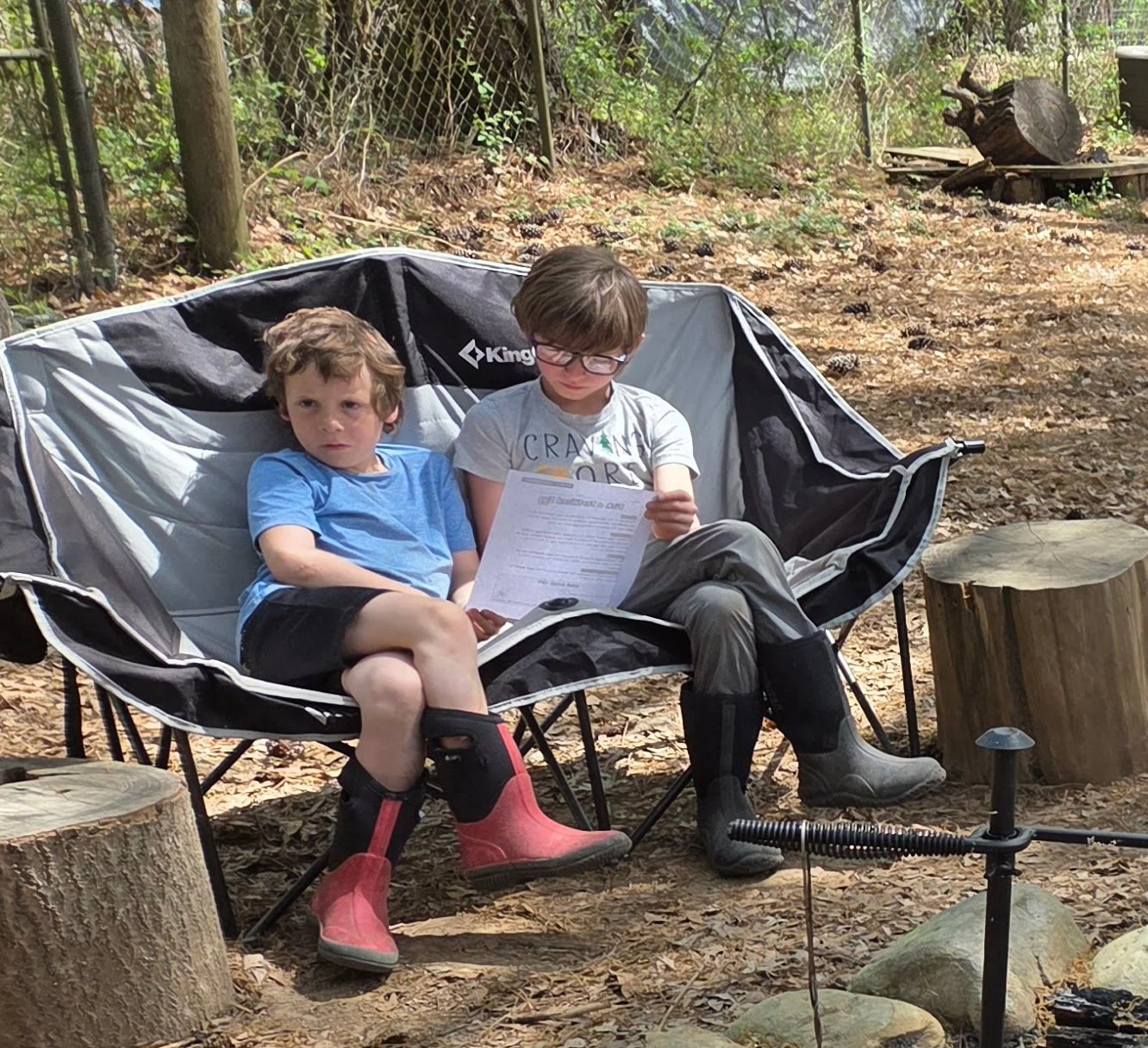 Two young boys sitting on a foldable camping chair outdoors, surrounded by trees and logs, one reading a paper and the other looking at the camera.