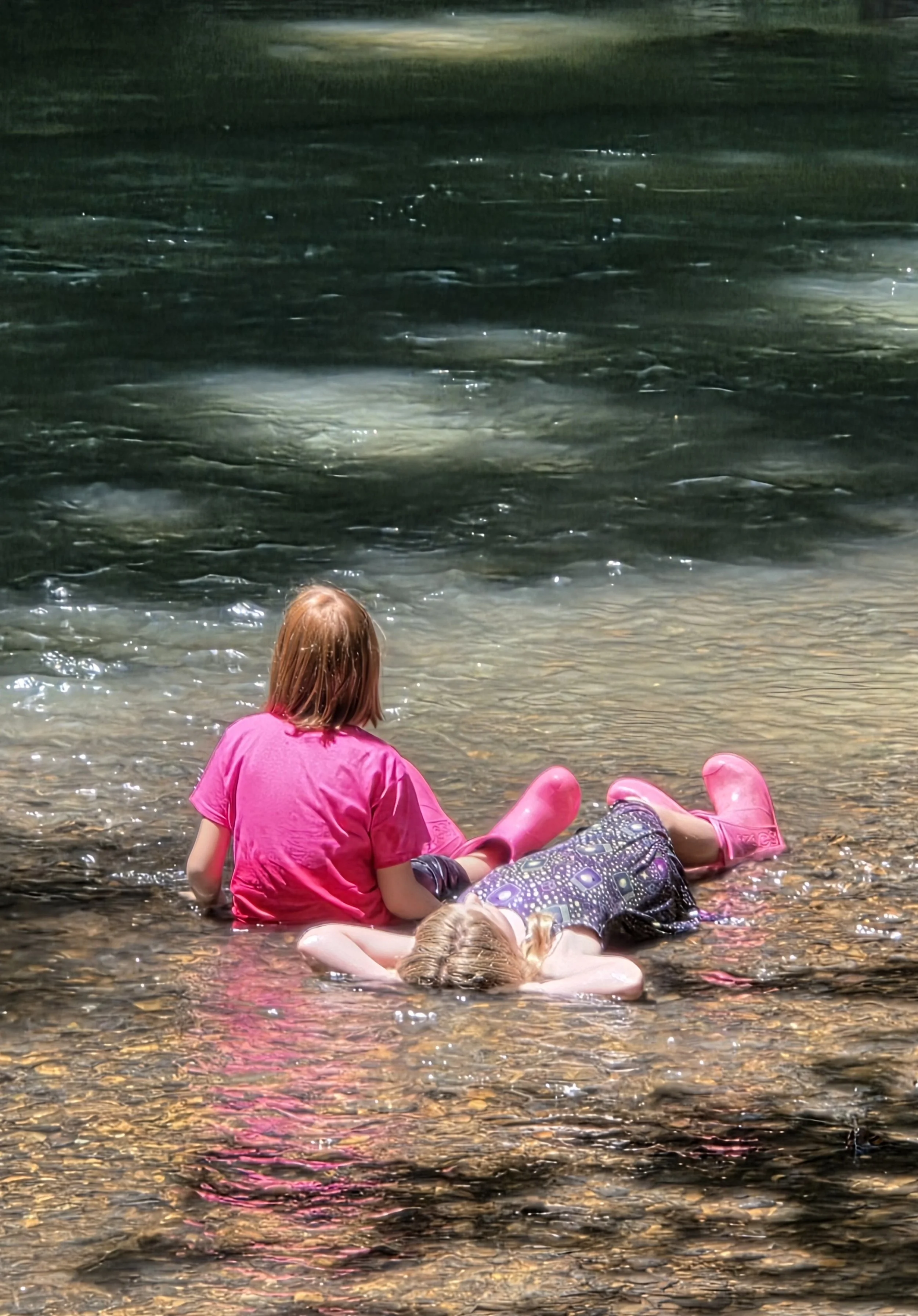 Children sitting and lying in shallow water on a beach at night, with one girl in a pink shirt and pink boots and another girl in a patterned dress and glasses.
