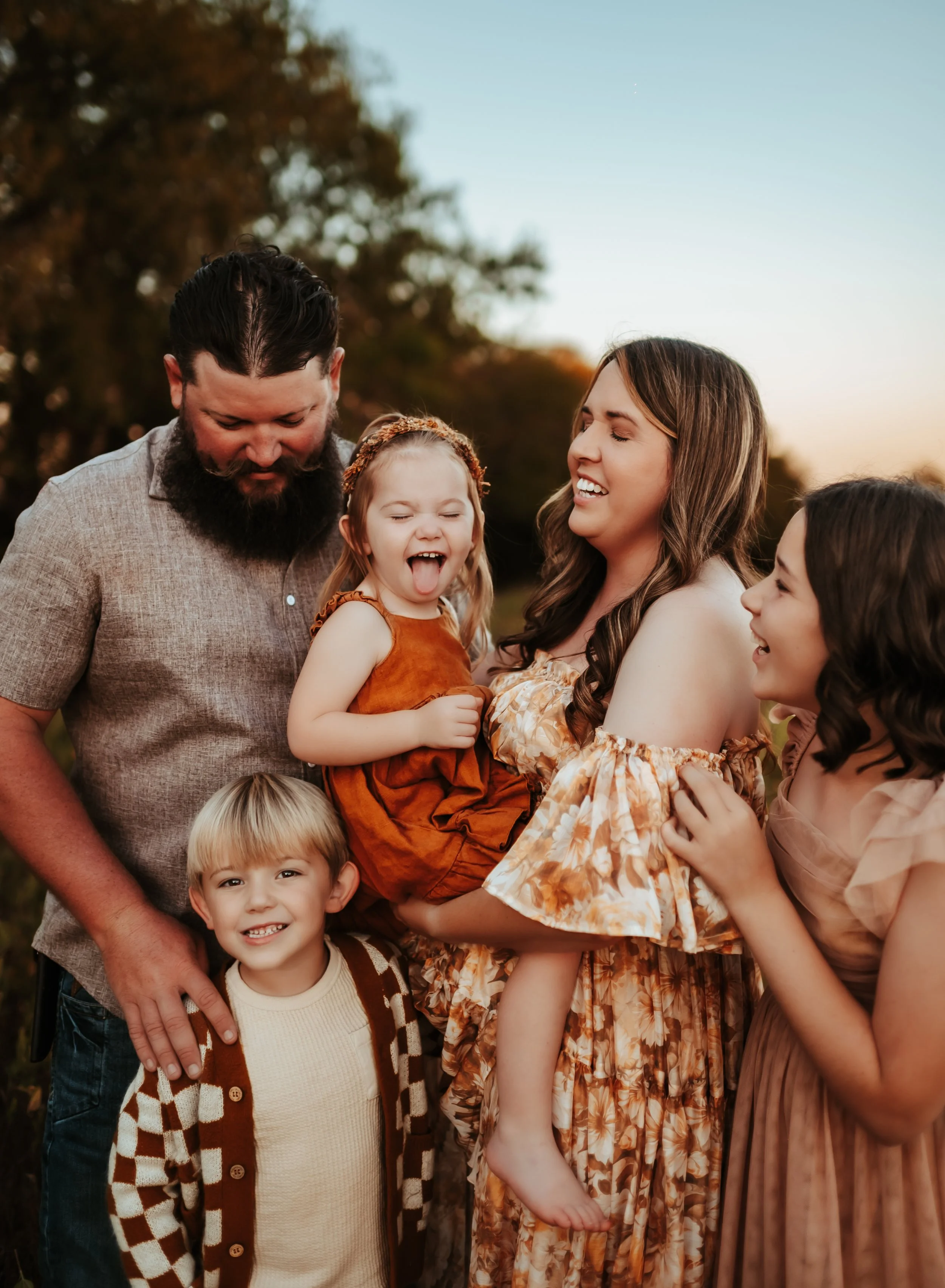 A family of six smiling and laughing outdoors at sunset with trees in the background, including two adults and four children.