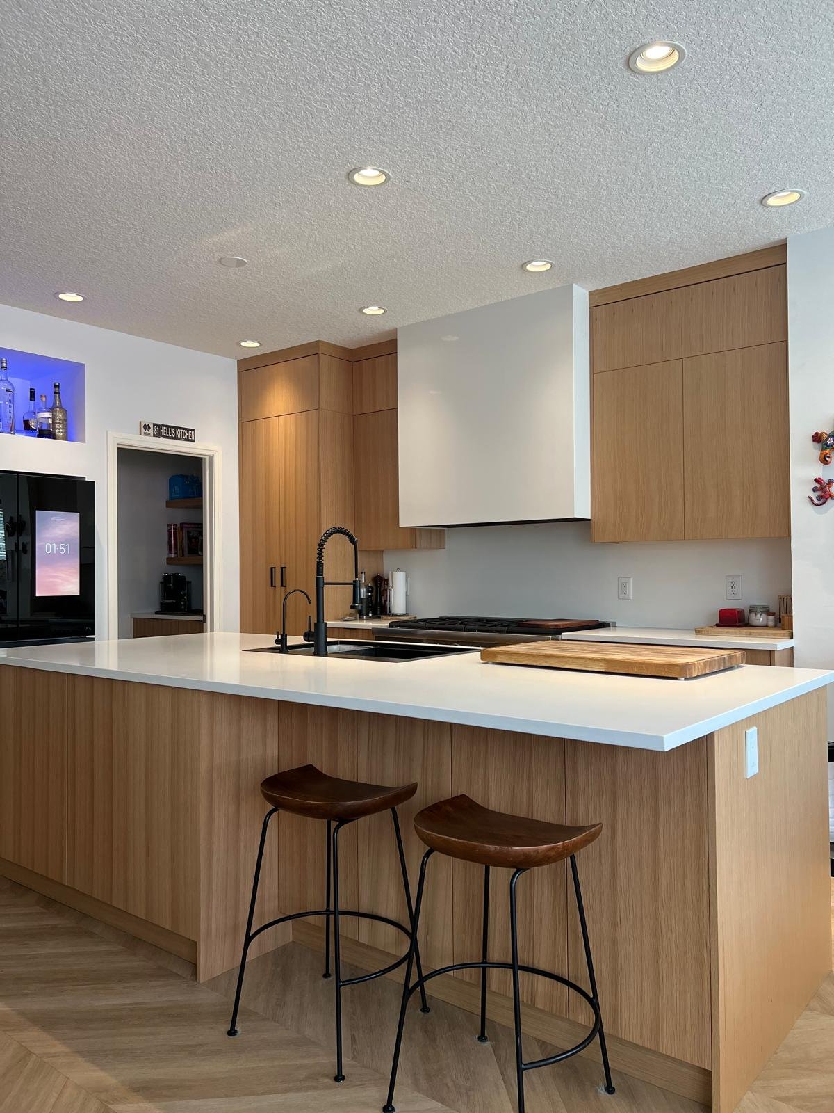 Modern kitchen with wooden cabinets, white countertop, black sink faucet, and two bar stools. There are small appliances and decor items on the counters, and a TV in the background.