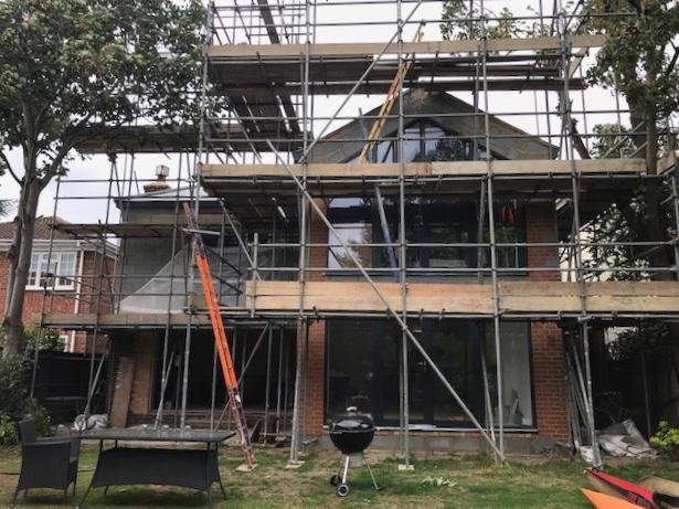 A two-story brick house under construction with scaffolding around it. There are trees and outdoor furniture in the yard, along with a small grill in front of the house.