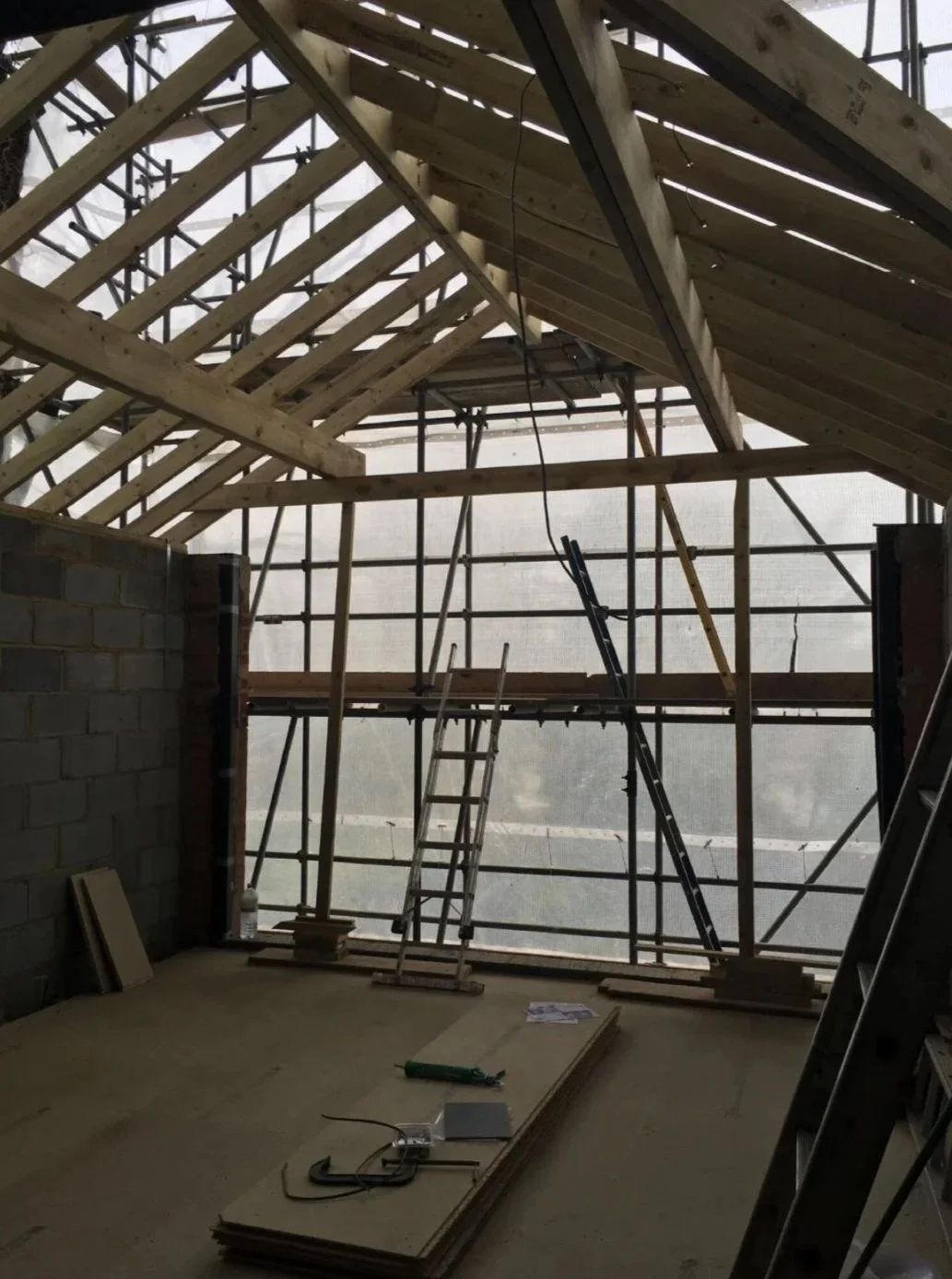 Construction site of a building with wooden framework for a roof and scaffolding inside. Some tools and materials are on the floor, and a ladder is leaning against the structure.
