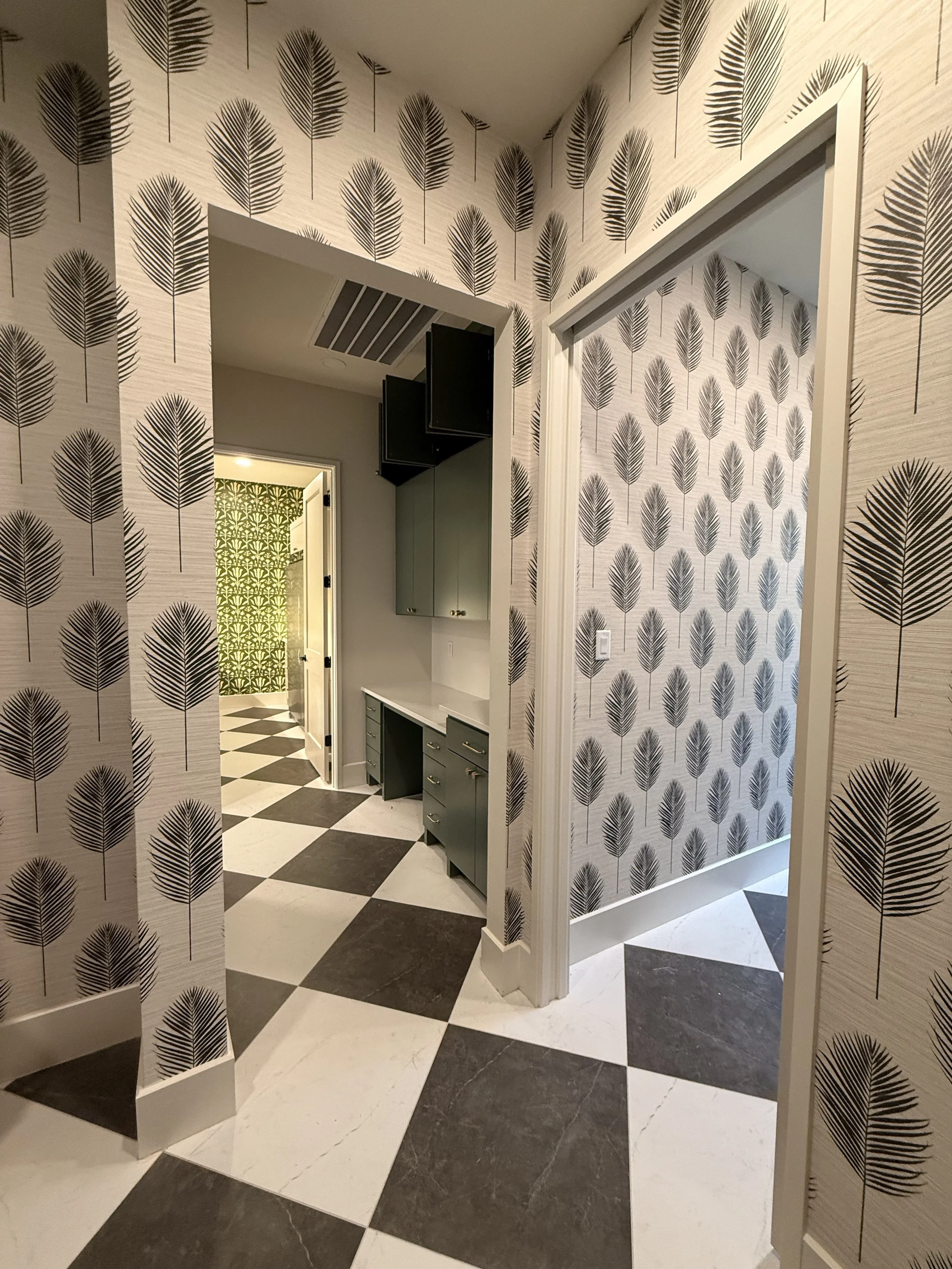 Interiors of a house with black and white checkered flooring, patterned wallpaper with black leaves, and a doorway leading to a room with green wallpaper with leaf pattern.
