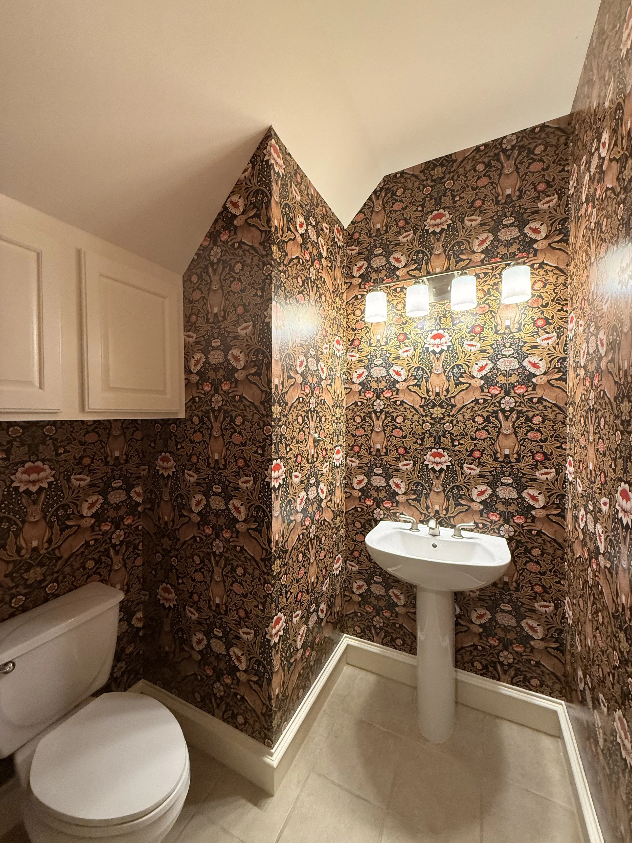Small bathroom with floral wallpaper, a toilet on the left, a white pedestal sink with a three-light fixture above, beige tile floor, and white cabinets.