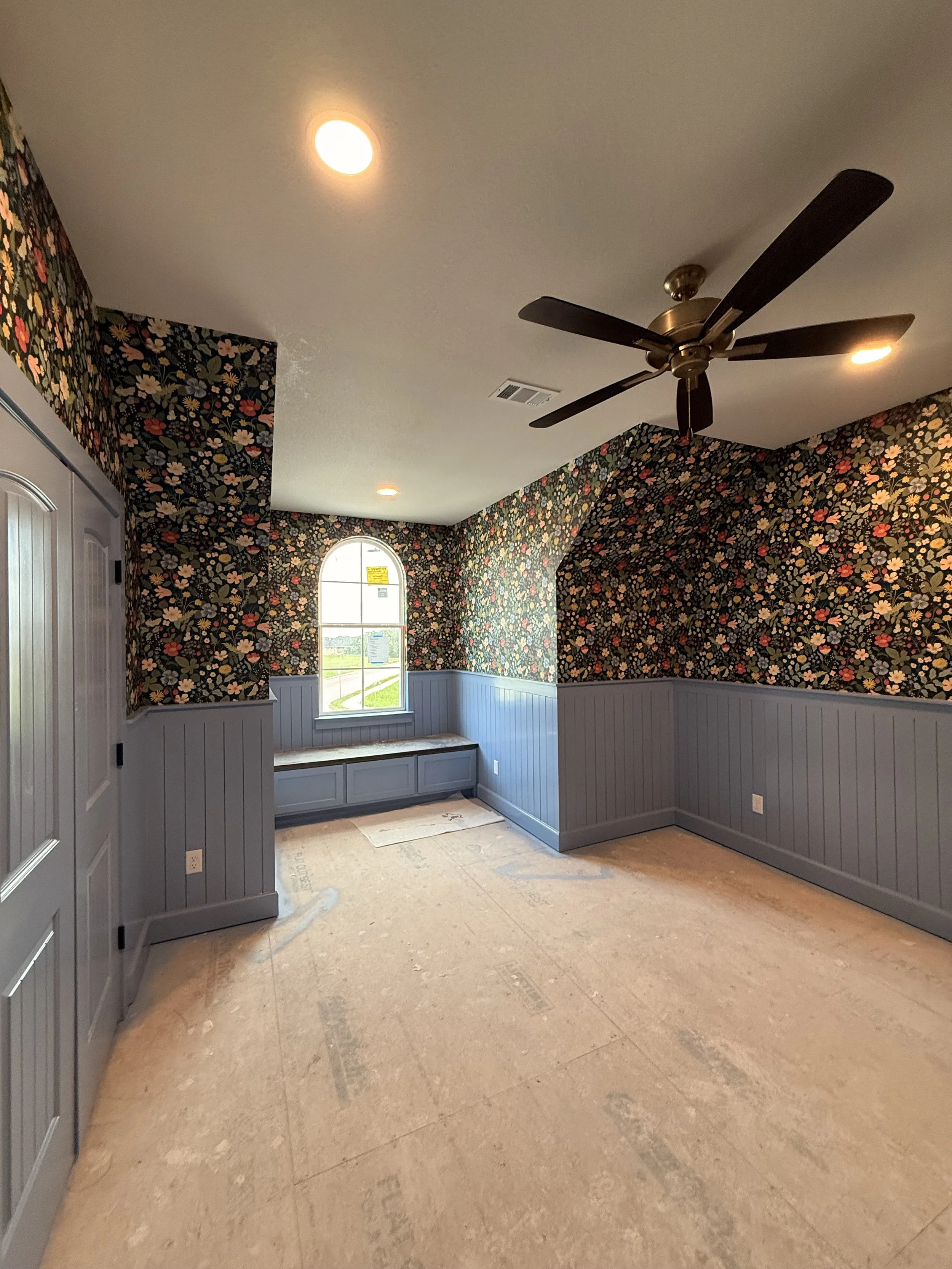Interior room with floral wallpaper, wooden paneling below, a window, ceiling fan, and unfinished flooring.