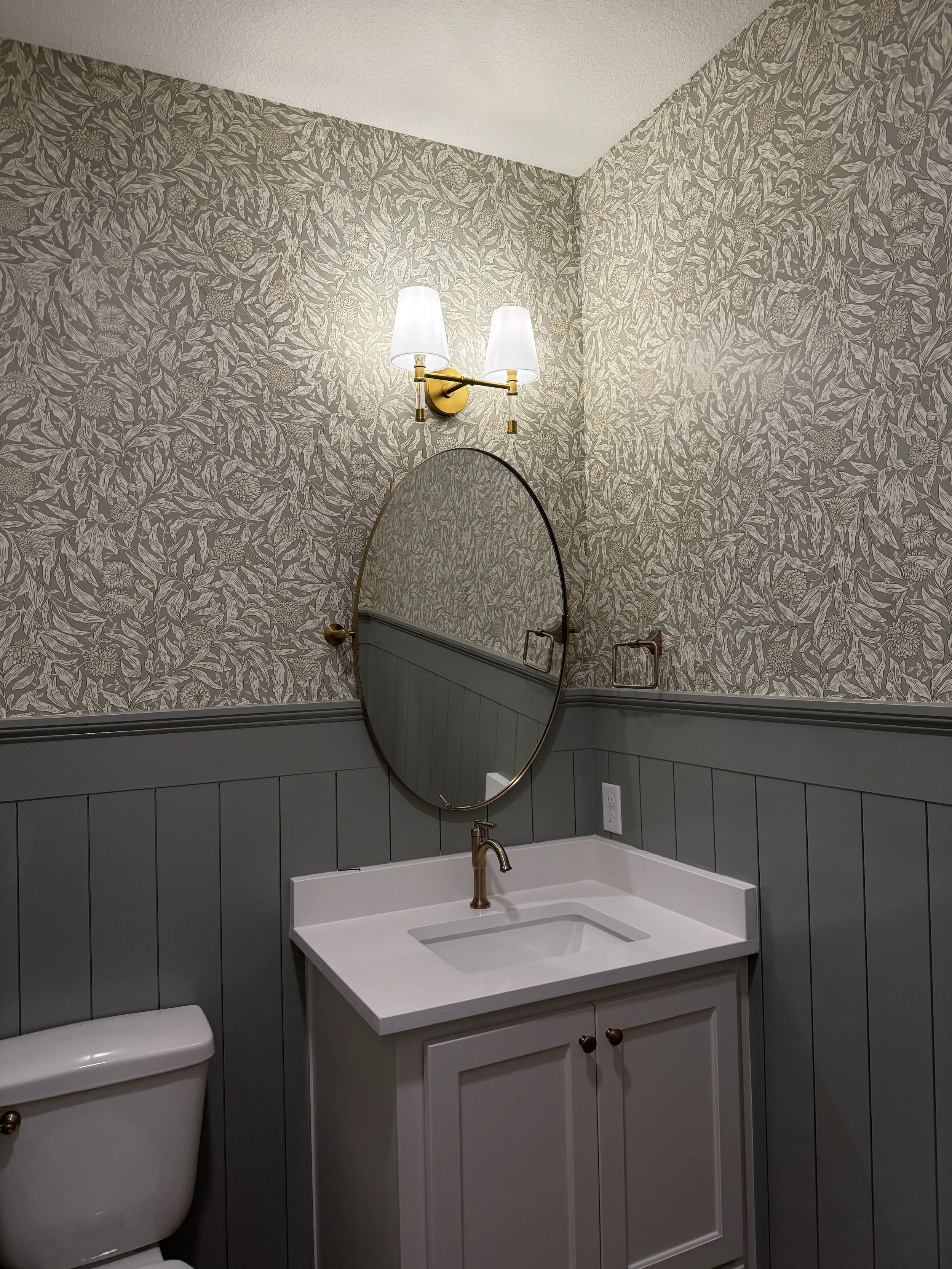Bathroom sink with a gold faucet, wall-mounted mirror with gold frame, wall-mounted light fixture with two white lampshades, wallpaper with floral pattern, and gray paneling below the wallpaper.