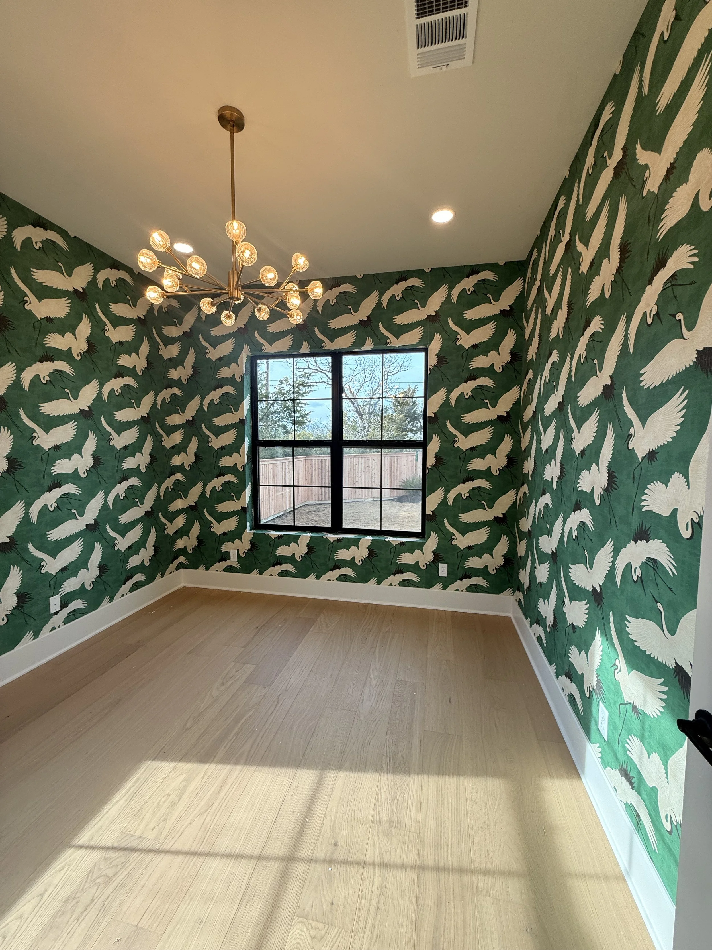 A room with green wallpaper featuring white cranes, a large window showing trees outside, and a modern chandelier with exposed bulbs hanging from the ceiling.
