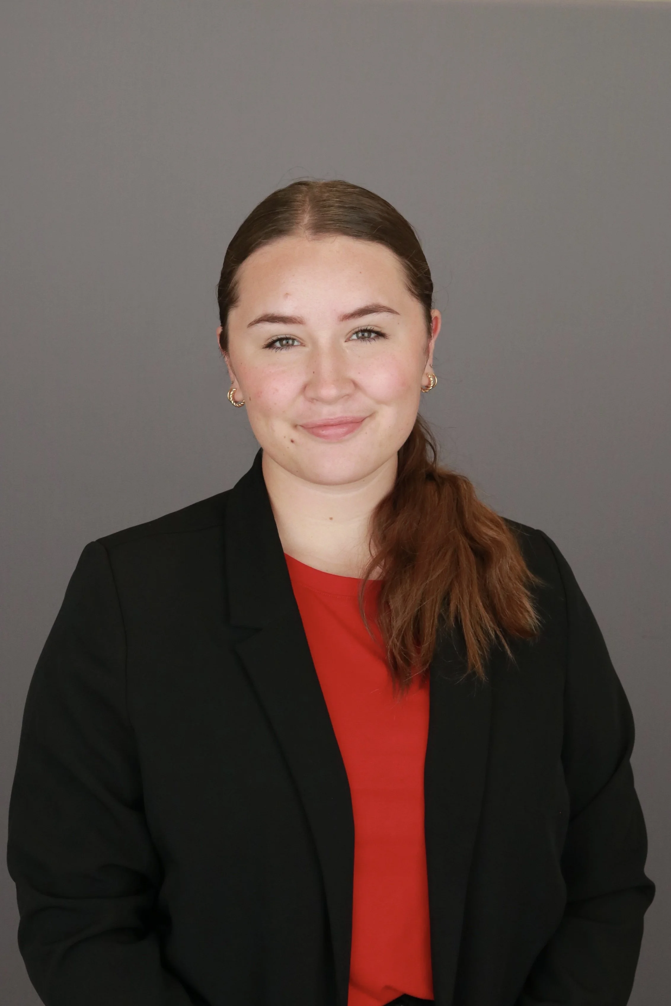A woman with brown hair, wearing a black blazer and a red shirt, standing against a gray background.