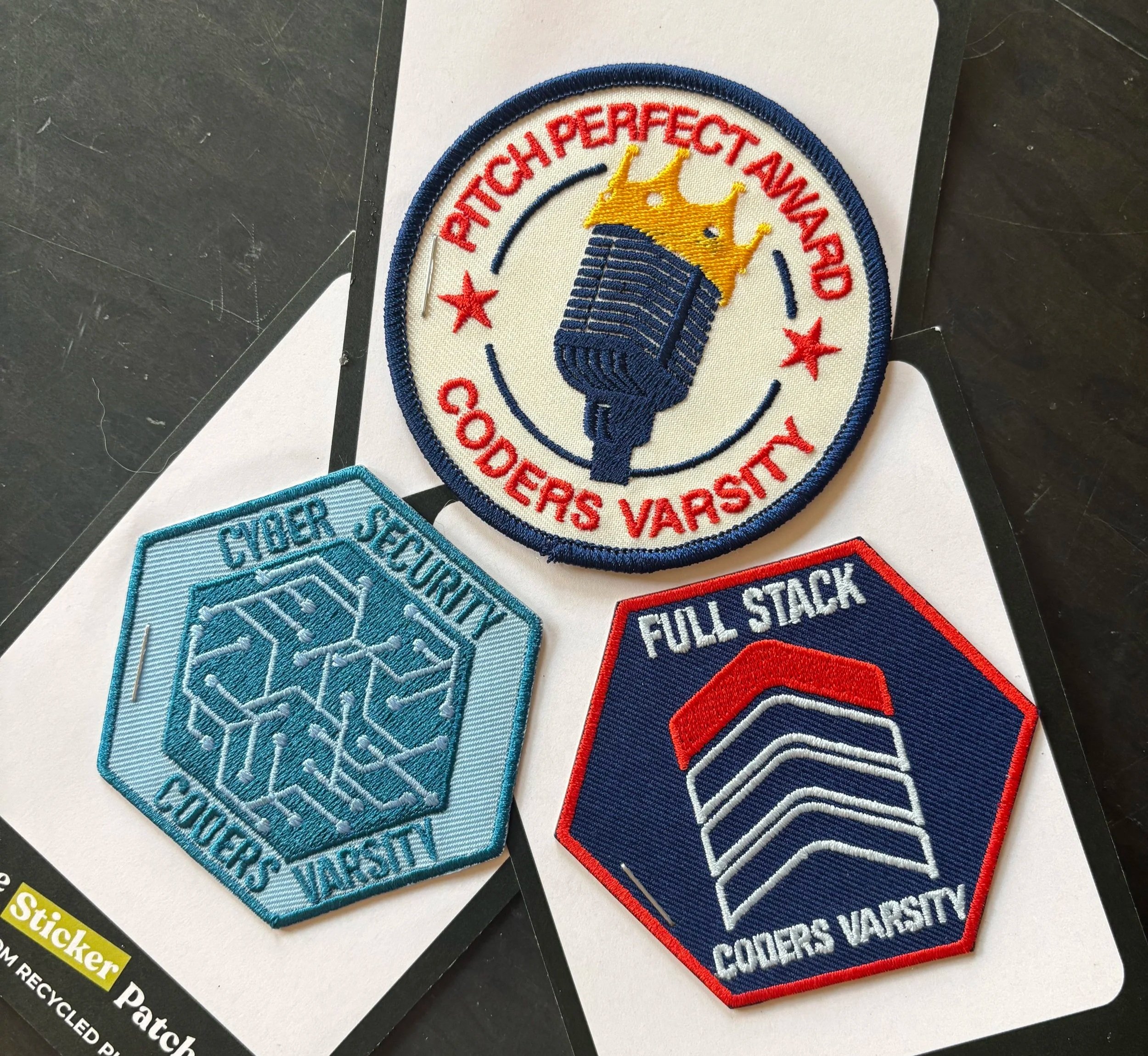 A set of embroidered patches of a microphone with a crown stating 'PITCH PERFECT AWARD' and 'CODERS VARSITY' around it, a stack of chevrons for 'FULL STACK', and circuit board designs in the shape of a hexagon for 'CYBER SECURITY'.