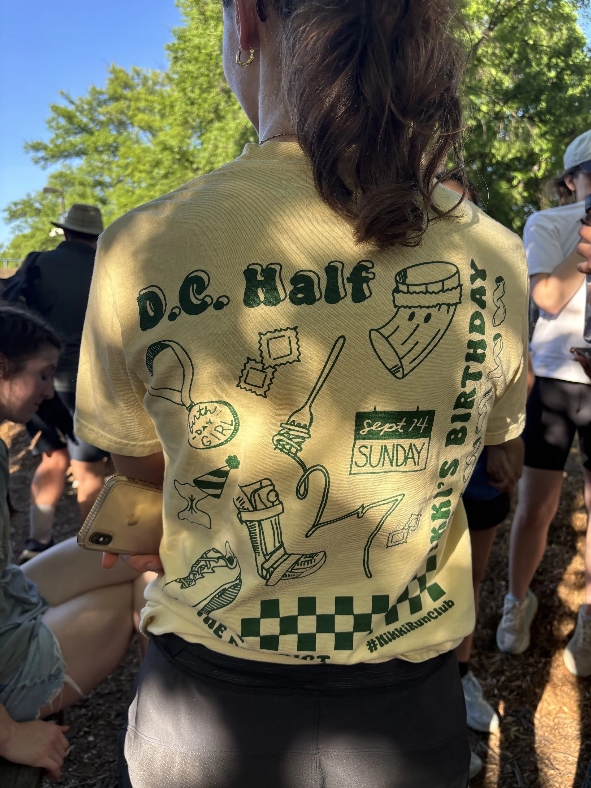 Back of a person wearing a yellow T-shirt with dark teal graphics and text, surrounded by other people outdoors with green trees and blue sky in the background.