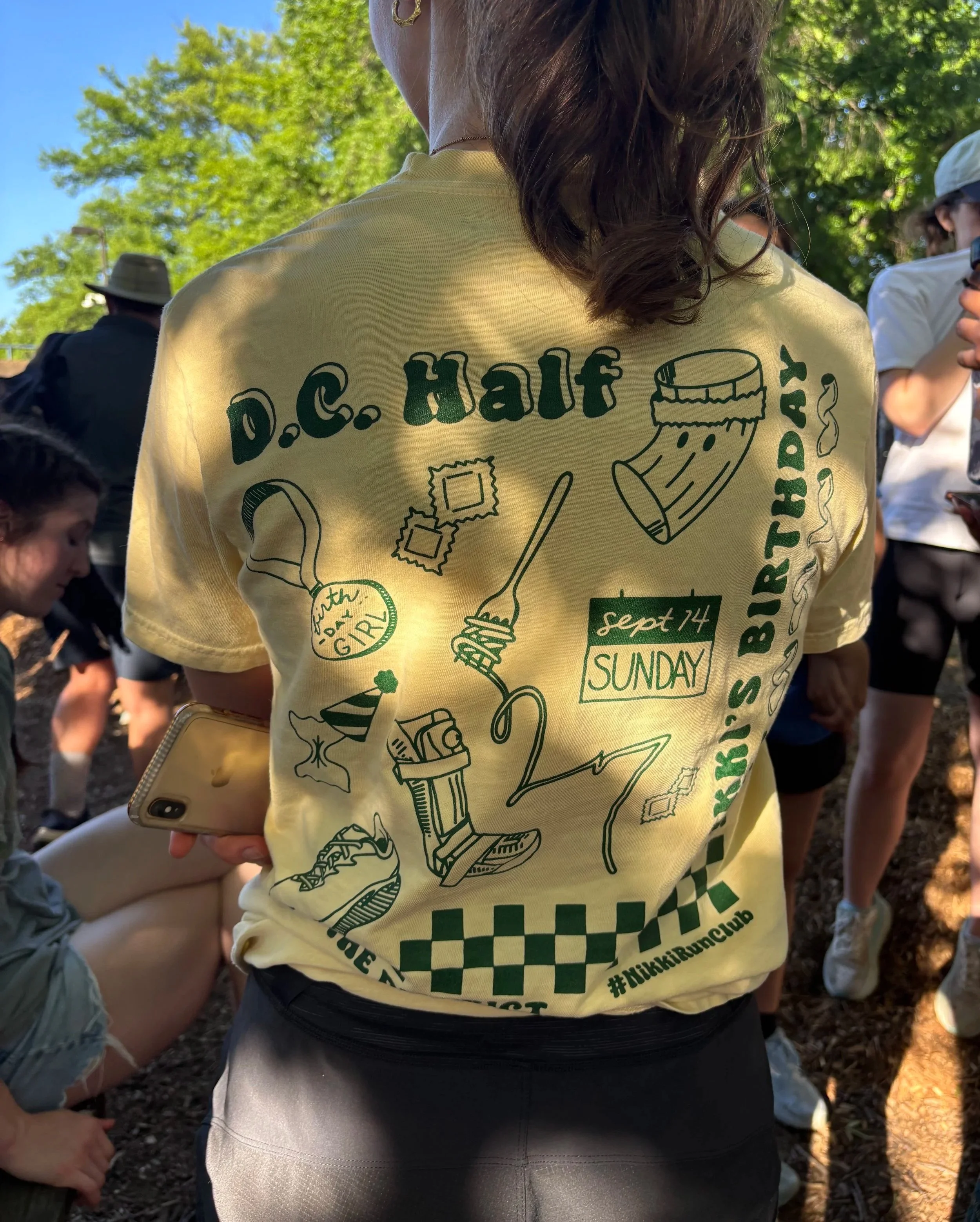Close-up of a person wearing a yellow t-shirt with graphic designs and text, with others gathered outdoors in the background, sunlight filtering through trees.