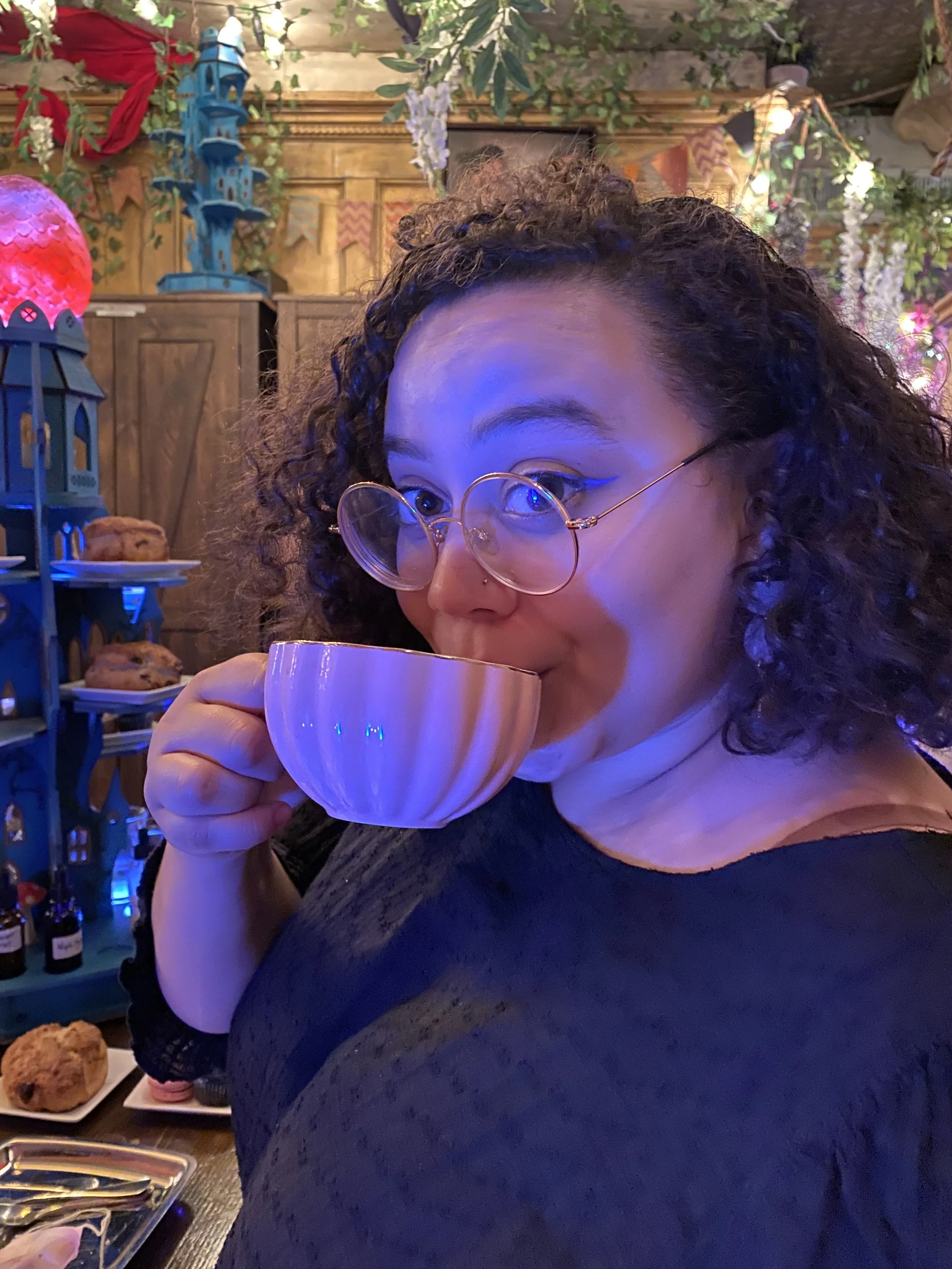 A woman with curly hair and glasses drinking from a pink cup in a cozy, decorated interior with plants, fairy lights, and shelves of baked goods and bottles.