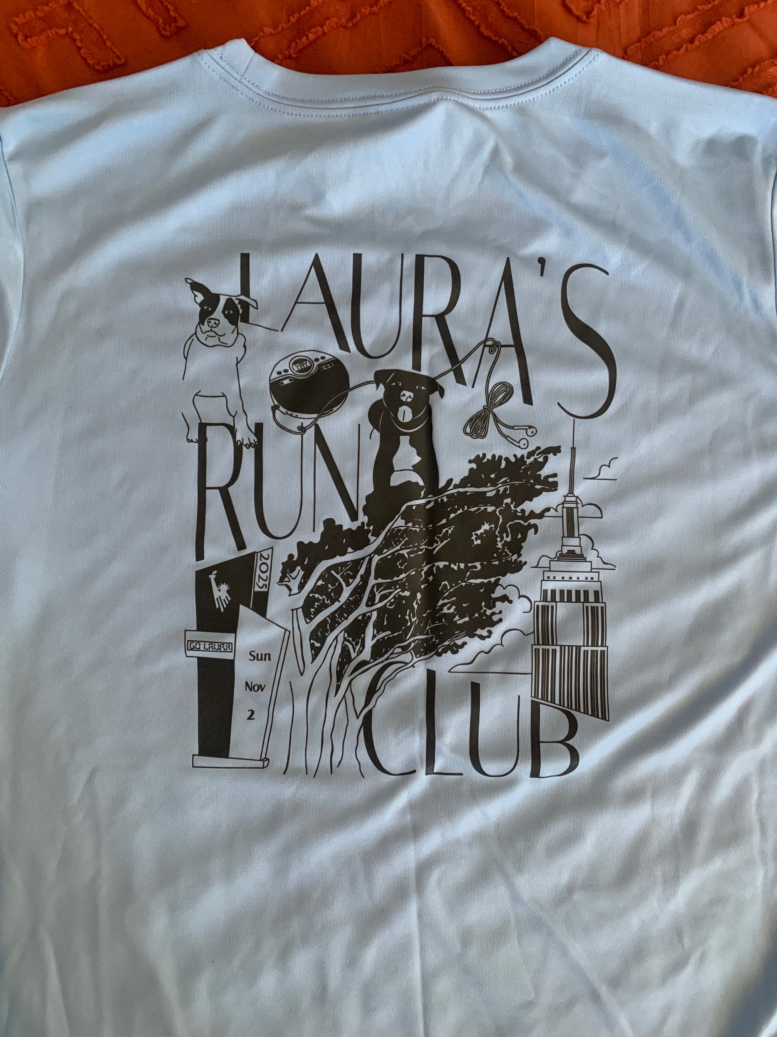 A light green T-shirt with a dark teal graphic design and text reading 'LAURA'S RUN CLUB'. The design includes a line drawing of a dog, a tree, a radio, the Empire State Building, and a street sign.