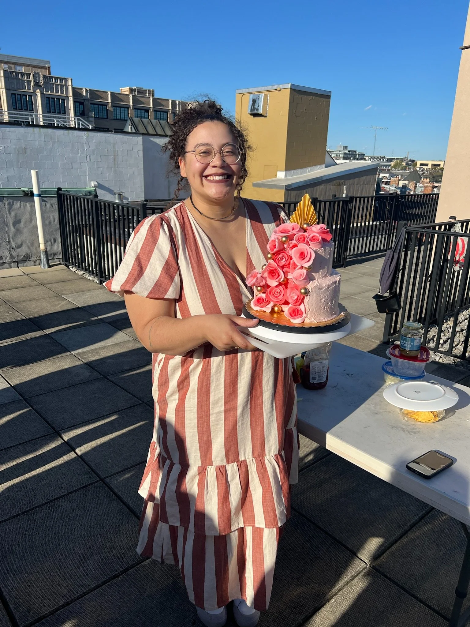 A woman with glasses and curly hair holding a pink cake decorated with pink roses and gold accents on a rooftop.