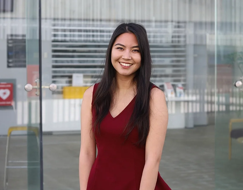 A young woman with long dark hair smiling and wearing a sleeveless red dress, standing in front of a glass wall with an urban background.