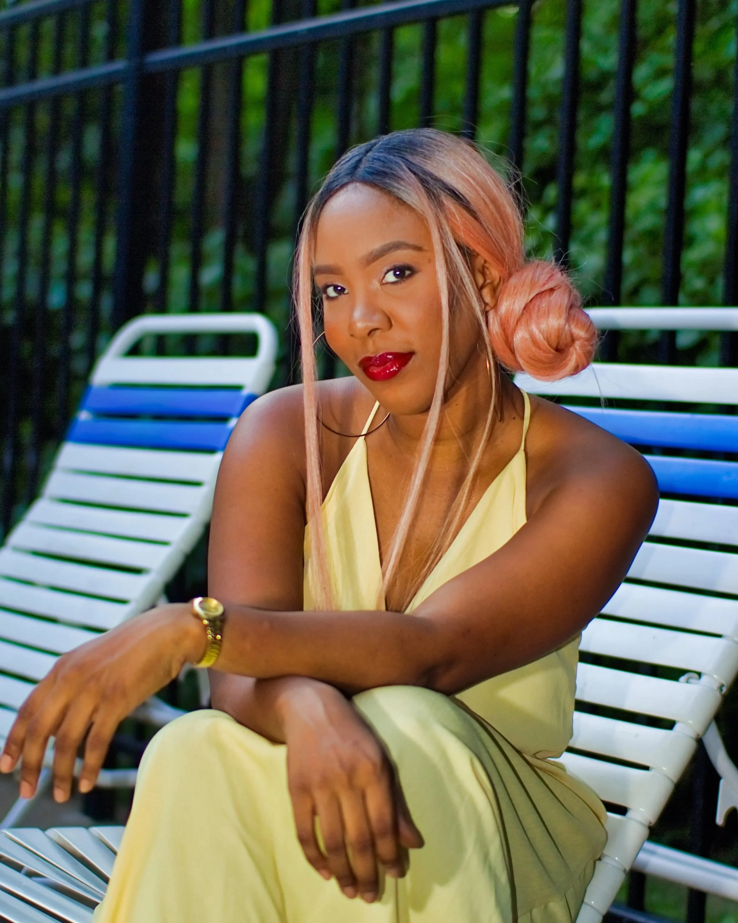 A woman with pinkish-blonde hair styled in a side bun, wearing red lipstick, gold hoop earrings, a yellow halter dress, and gold jewelry, sits outside on a white park bench with a black metal fence and green trees in the background.