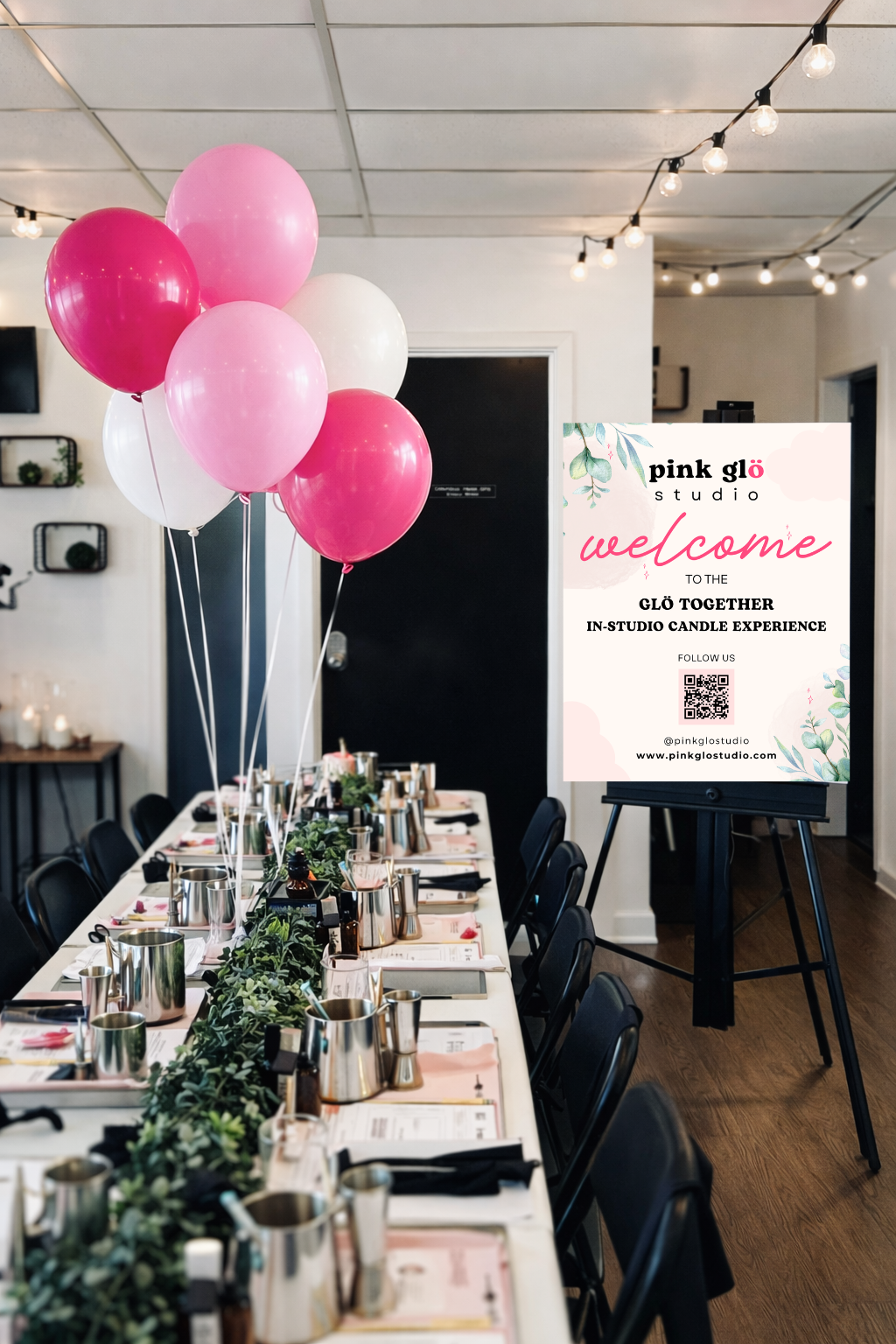 A decorated event space with a long table set for a candle-making experience, adorned with pink, white, and red balloons, greenery runner, and place settings. A welcome sign on an easel displays information about the Pink Glö Studio's in-studio candle experience.