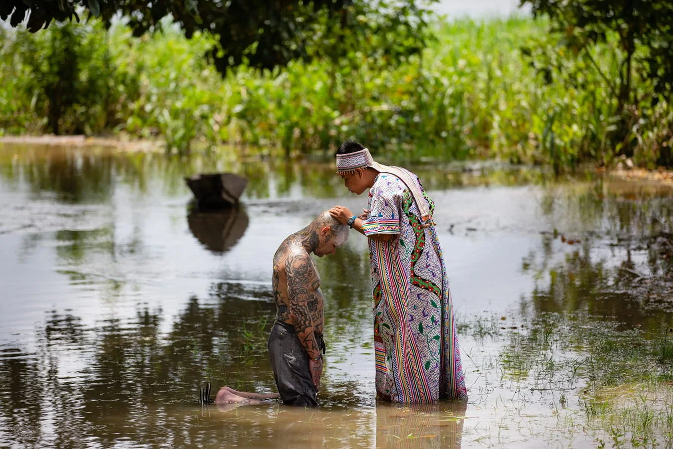 A person with tattoos kneeling in water, head bowed, while another person in traditional indigenous attire pours water over their head in a lush outdoor setting.