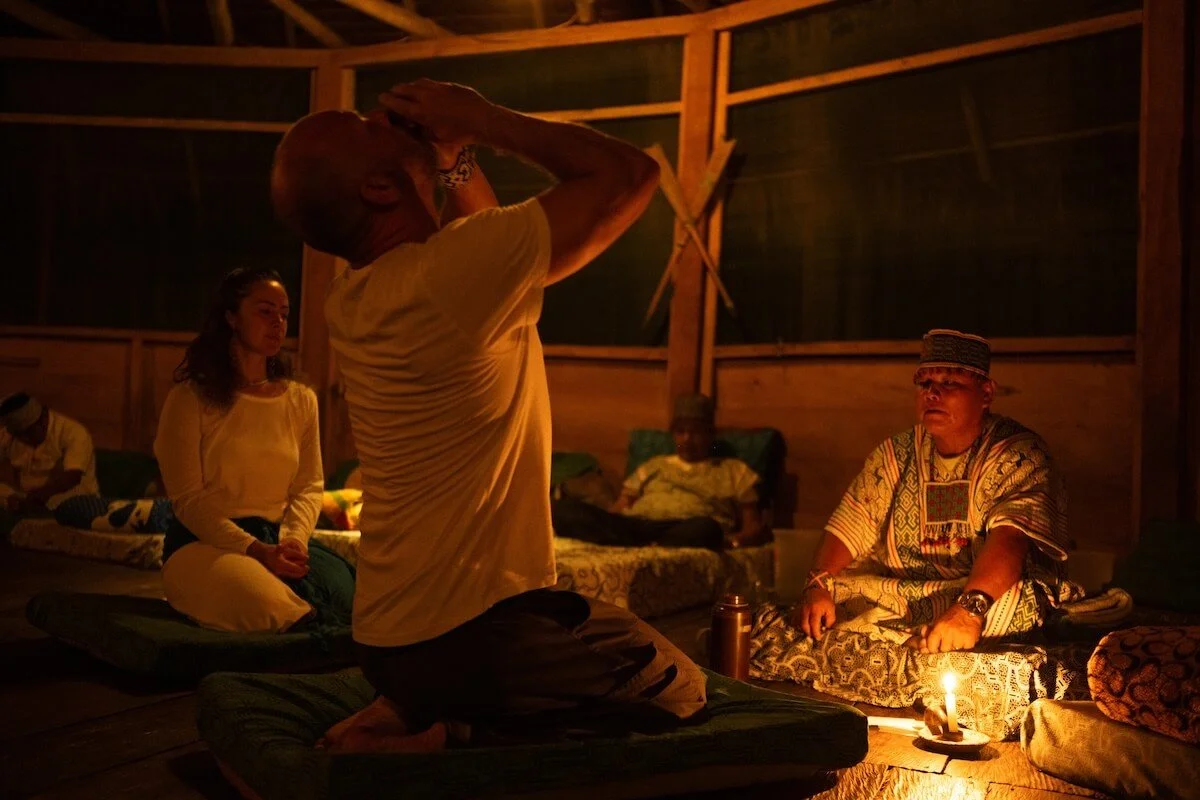 Group of people sitting on layered mats inside a wooden structure, gathered around a lit candle. One person is kneeling with hands over head, while others sit peacefully, some with eyes closed.