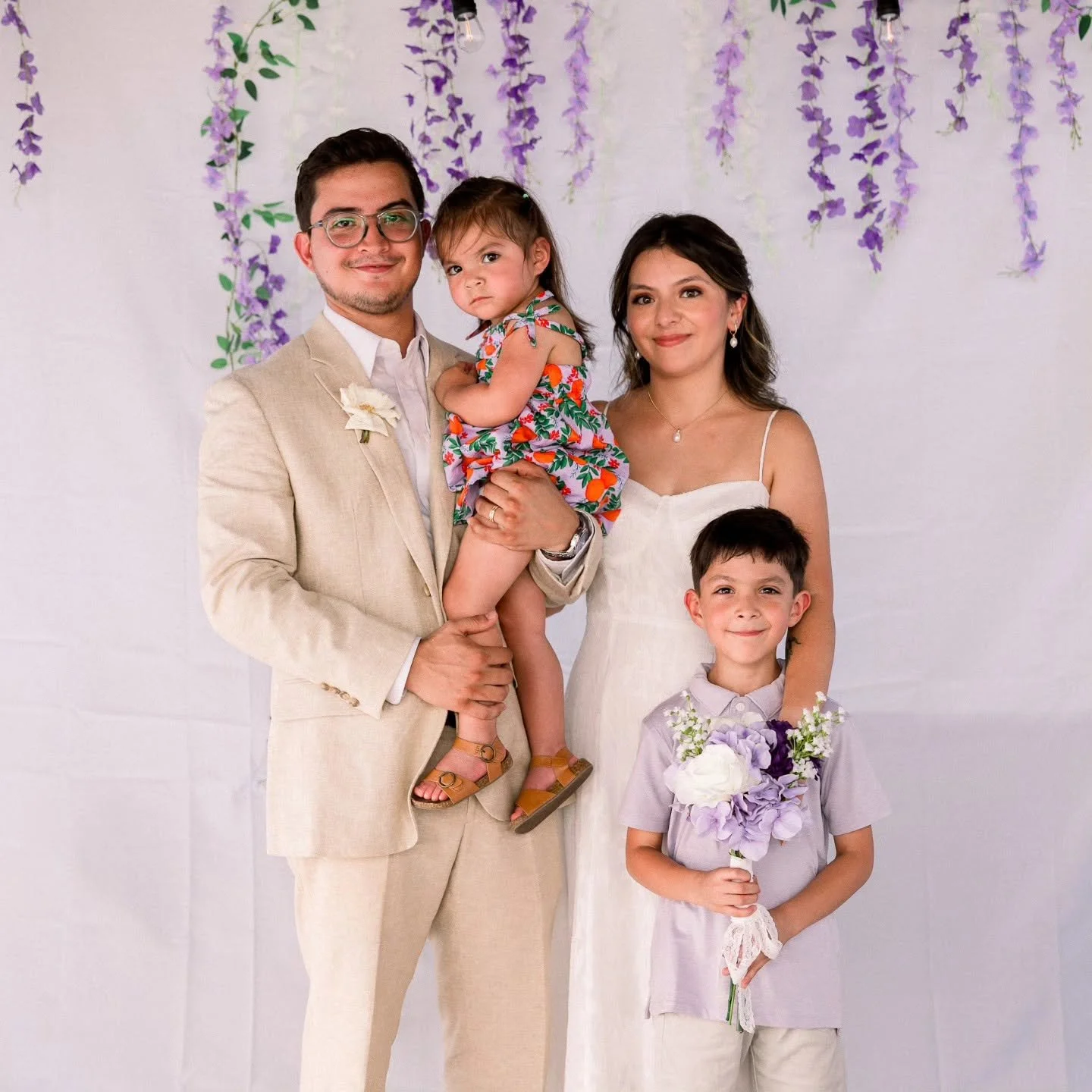 A family of five with two adults and three children posing in front of a floral backdrop at a celebration or wedding. The man is wearing a light beige suit, white shirt, and glasses. The woman is in a white dress with shoulder straps and pearl earrings. The girl is being held by the man, wearing a colorful floral dress. The young boy is holding a bouquet of flowers, wearing a light purple shirt and beige pants. The family appears happy and dressed nicely for the occasion.