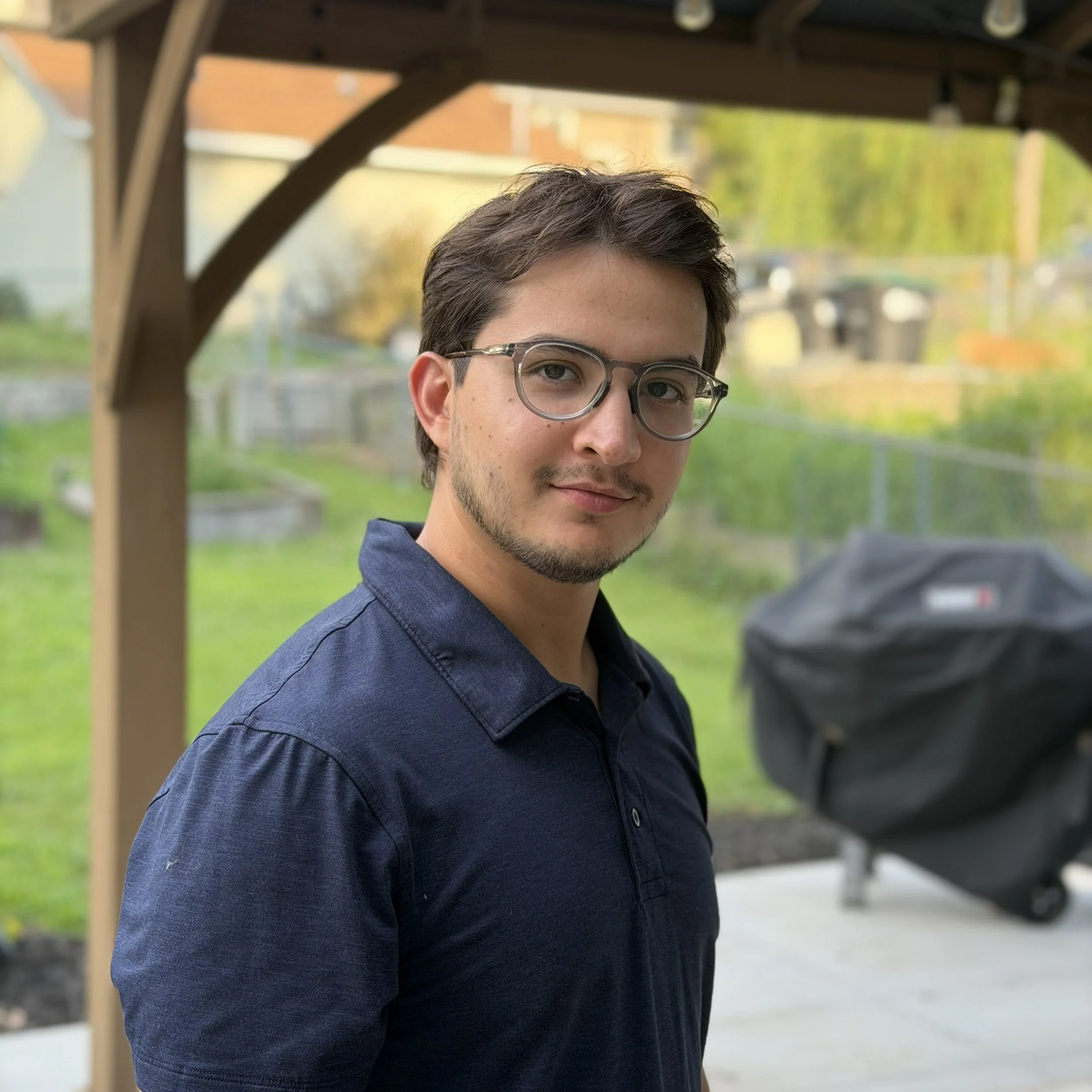 Close-up of a young man with short dark hair, wearing glasses and a navy blue collared shirt, standing outdoors with a blurred background of residential houses, greenery, and a playground structure.