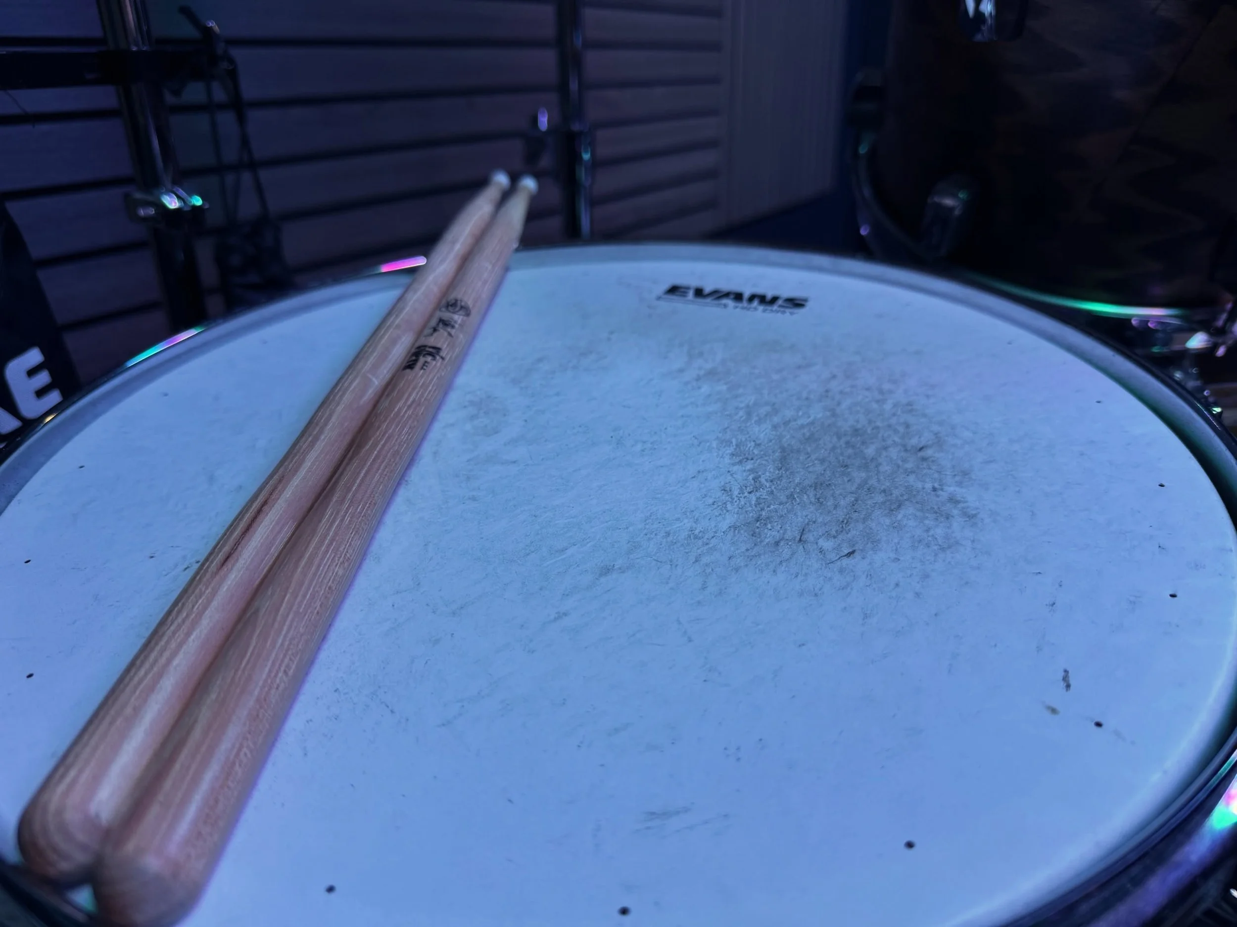 Close-up of a drum with drumsticks resting on it, against a dark background with a metal rack to the side.