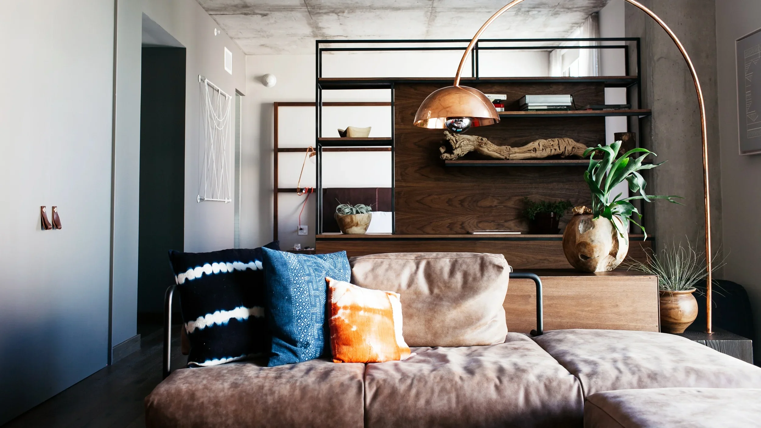 Modern living room with a sectional sofa, colorful pillows, wood shelving, plants, and an arched copper floor lamp