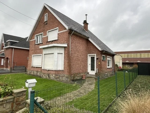 Red brick house with gray slate roof, fenced front yard, and neighboring buildings under an overcast sky