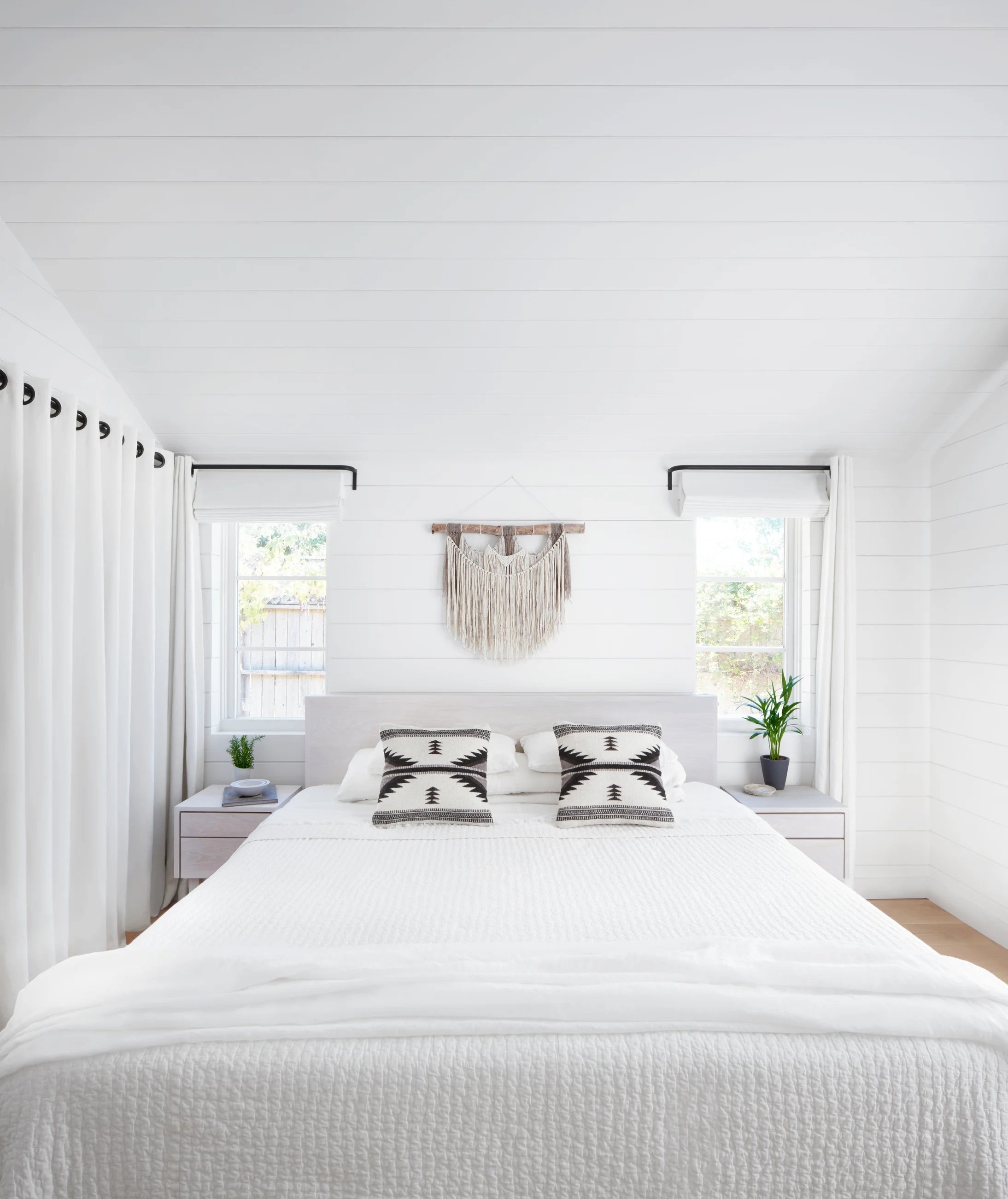 Bright white bedroom with a neatly made bed, patterned black-and-white pillows, wall hanging, windows, curtains, and potted plants