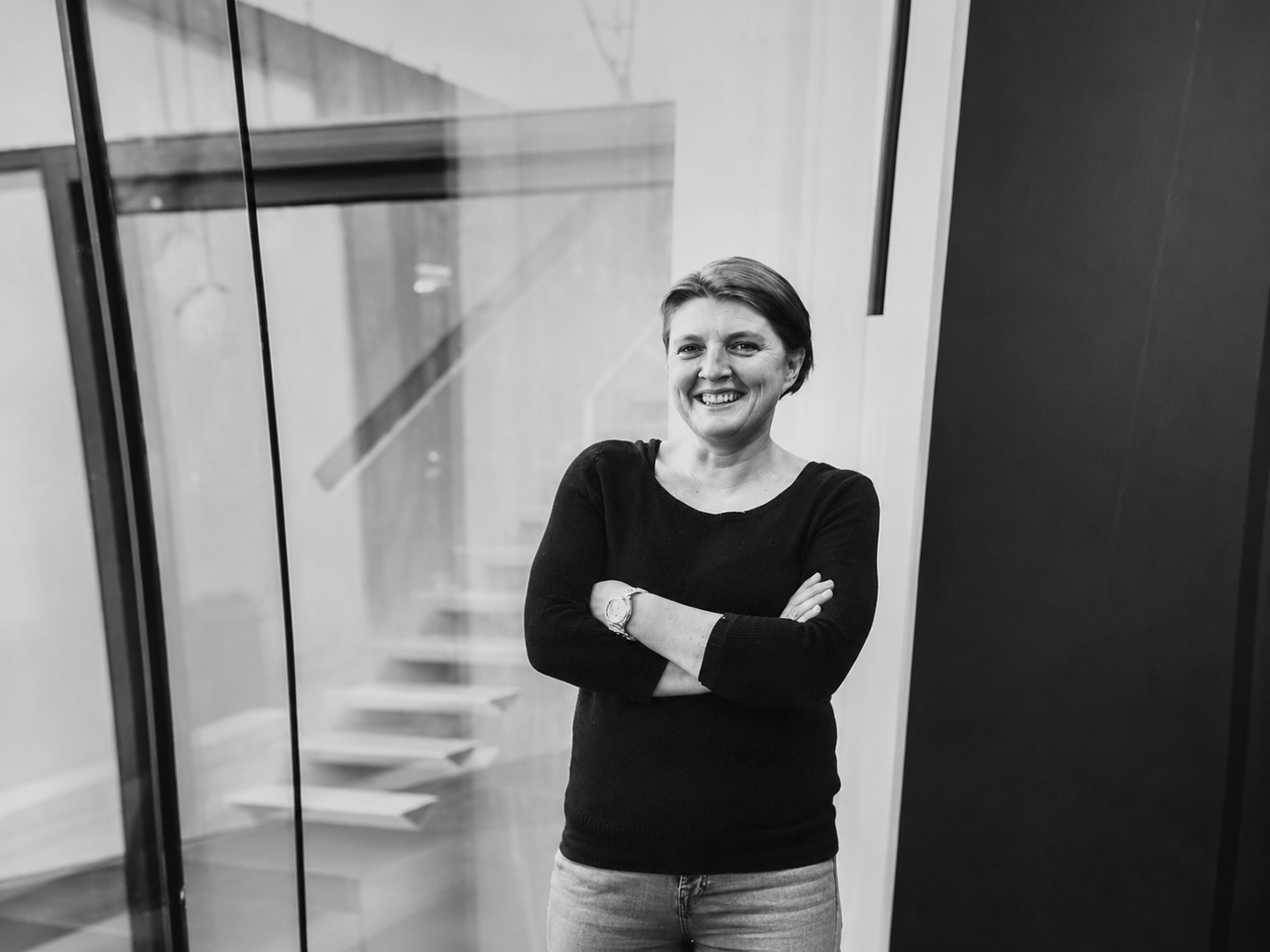 Smiling woman with crossed arms standing in a modern office beside a glass staircase