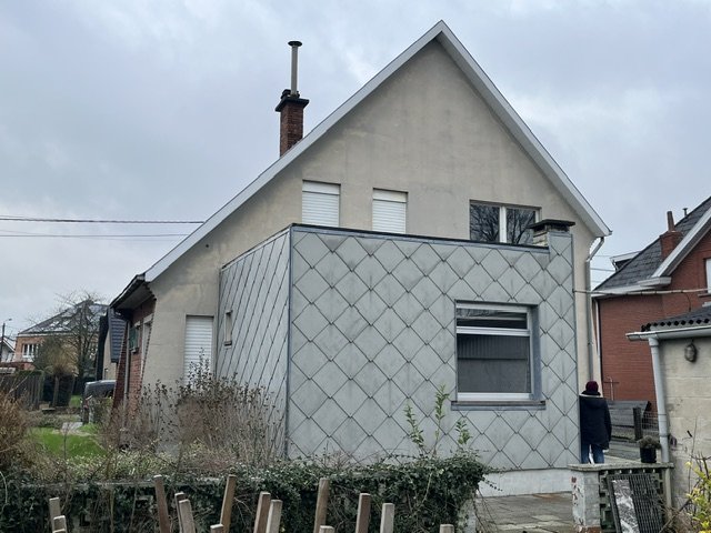 Small European house with patterned gray facade, steep roof, chimney, and front garden under an overcast sky