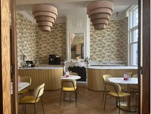 Elegant cafe interior with scalloped wallpaper, tiered pendant lights, marble fireplace, curved wood counters, and small dining tables.