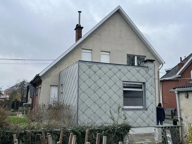 Small gray stucco house with diamond-patterned facade, steep gabled roof, and brick chimney on an overcast residential street