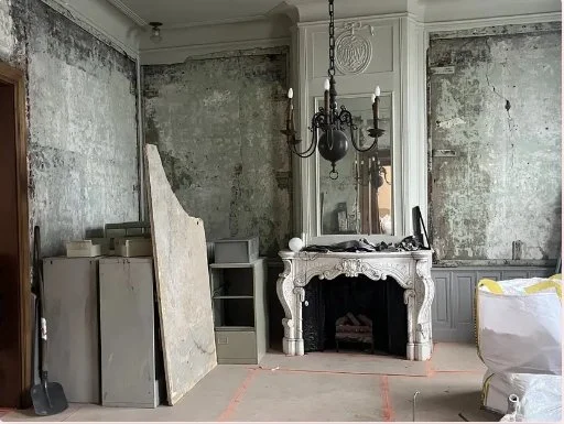Ornate white fireplace and chandelier in a distressed, partially renovated room with exposed plaster walls and construction materials