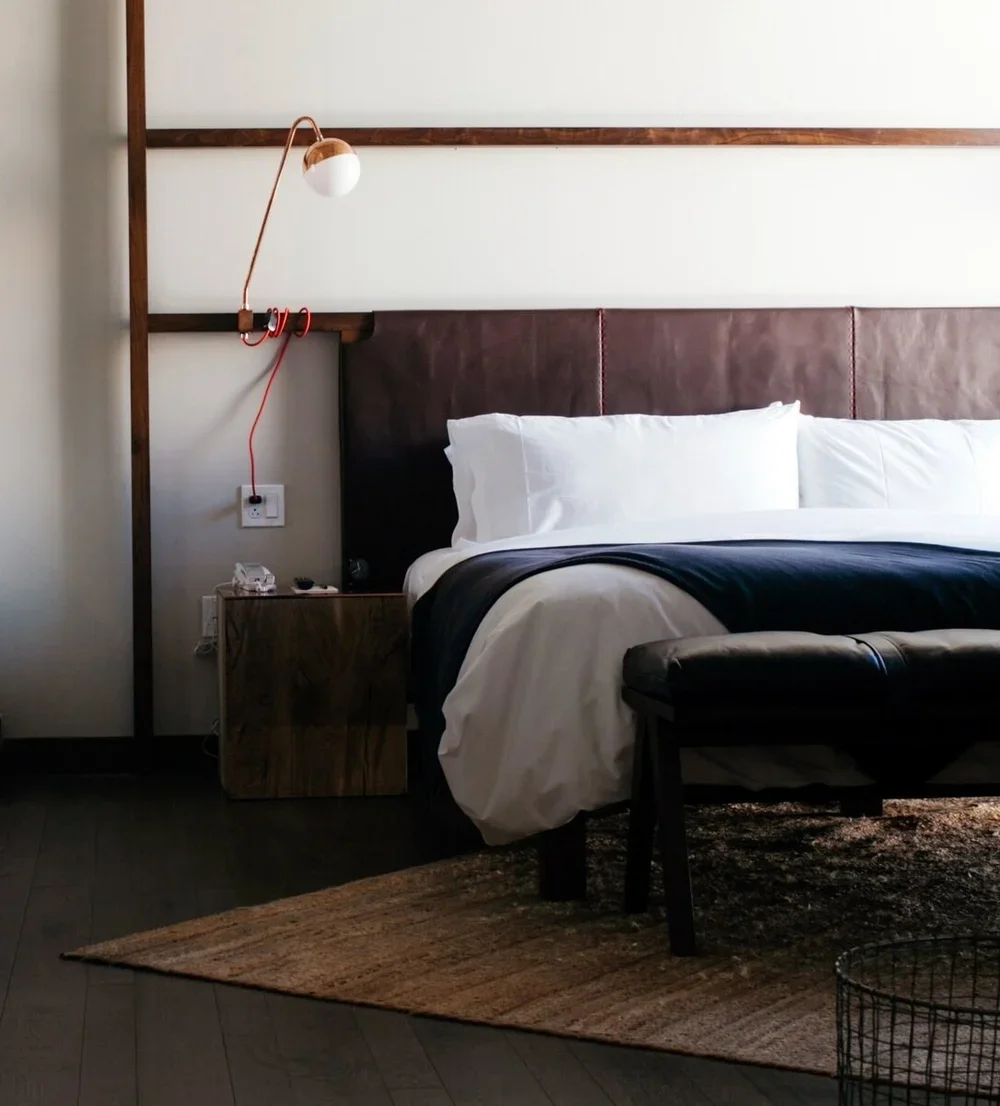 Modern bedroom with a white bed, dark leather headboard, wall-mounted lamp, wooden nightstand, and leather bench