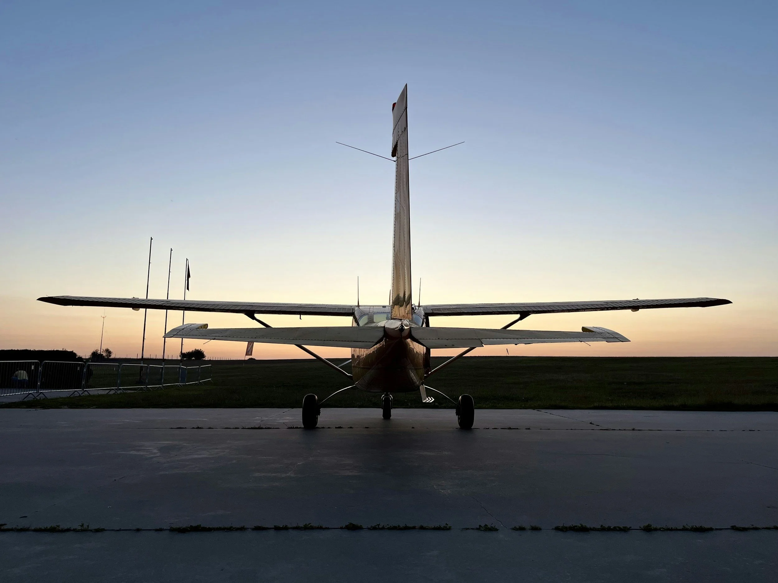 A small airplane parked on a tarmac at sunset with a clear sky and a few flags in the background.