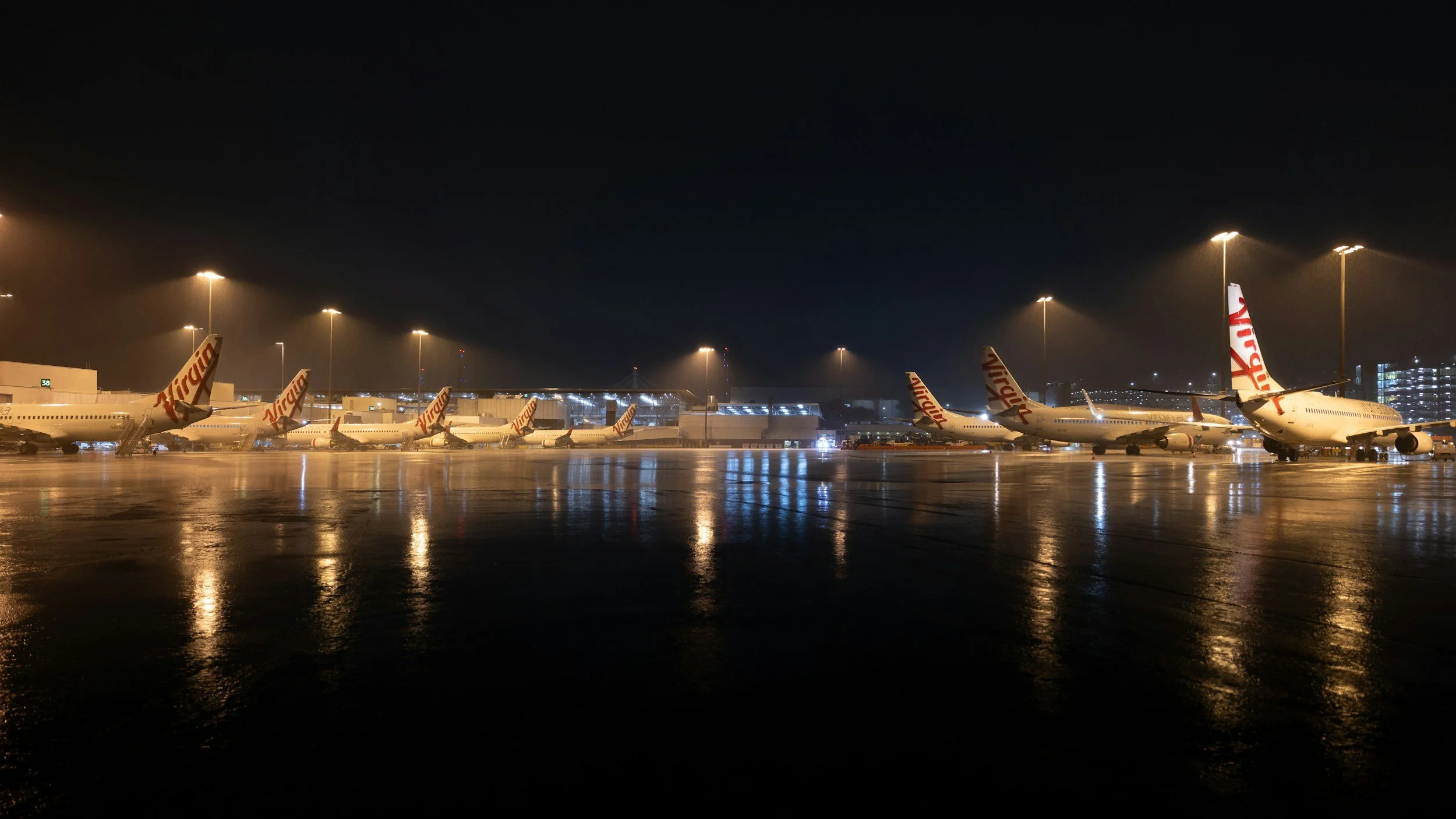 Nighttime view of commercial airliners parked at an airport tarmac with reflections on wet ground.