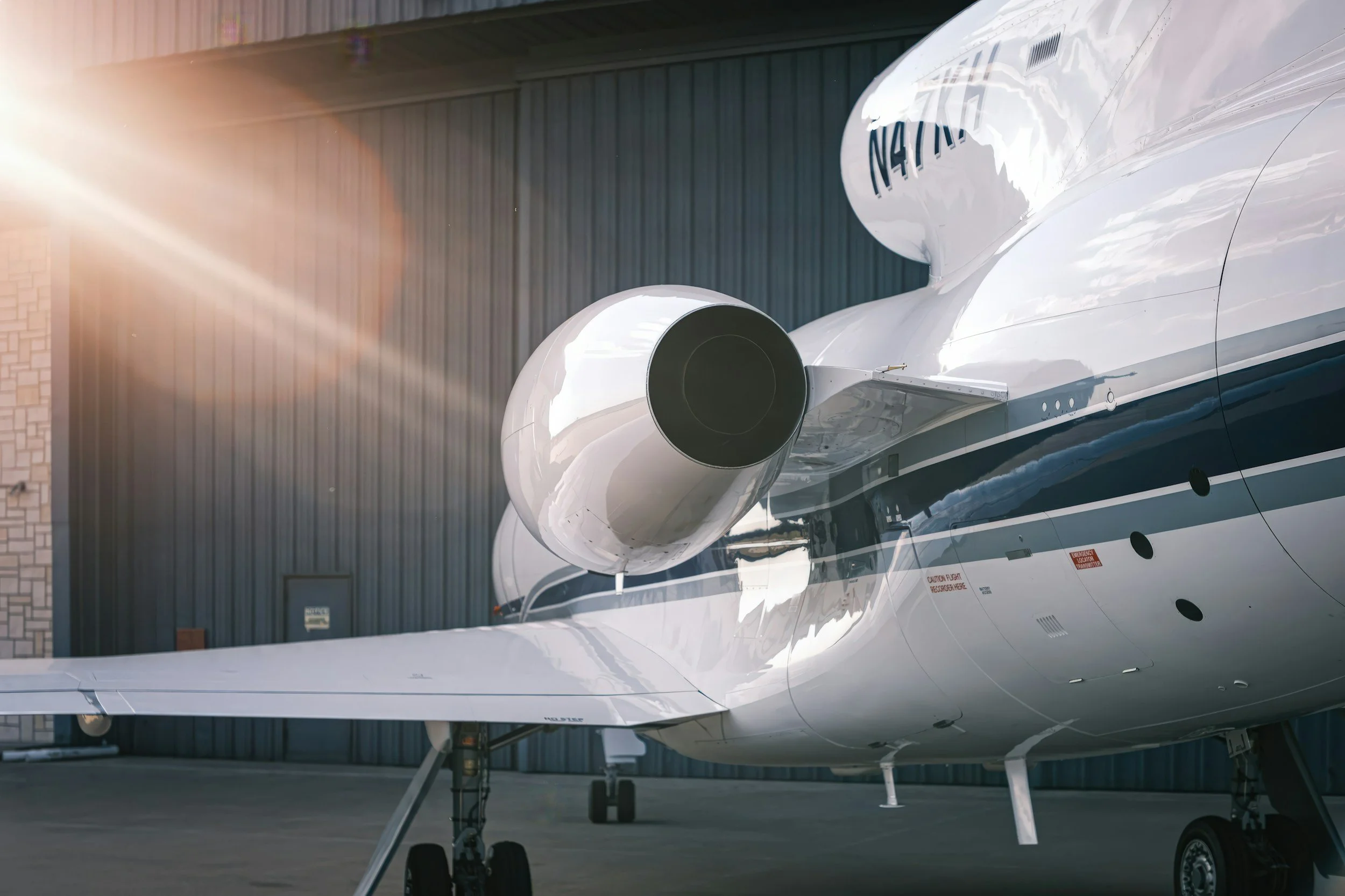 Close-up of the nose section and jet engine of a white private airplane, parked outside a hangar with sunlight shining.